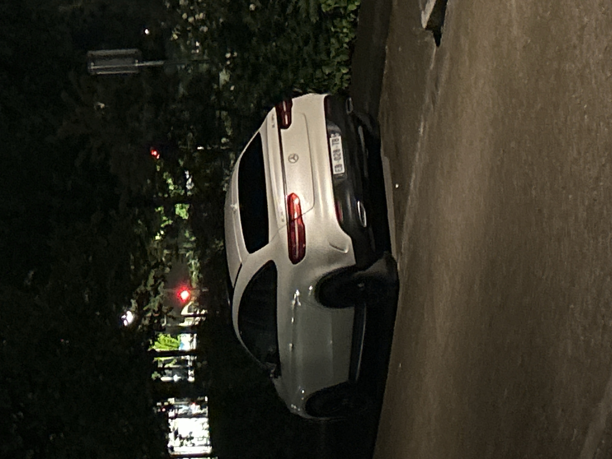 The image shows a white Mercedes-Benz parked on a street in Strasbourg, France at night. The license plate number is EB-028-TB. The vehicle is near a curb and foliage. In the background, there's a traffic light and illuminated buildings.
