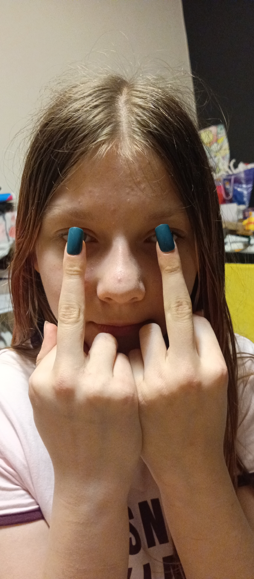 A close-up photograph depicts a young person with light brown hair, positioned indoors against a cluttered background with a yellow object. The individual is wearing a light pink shirt. Their eyes are partially obscured by their middle fingers, which are extended upwards. The fingernails are painted a dark teal color. The person's hands are positioned near their face, and their fingers are interlaced. The location is an indoor space. There are no indicators of city or any particular location.