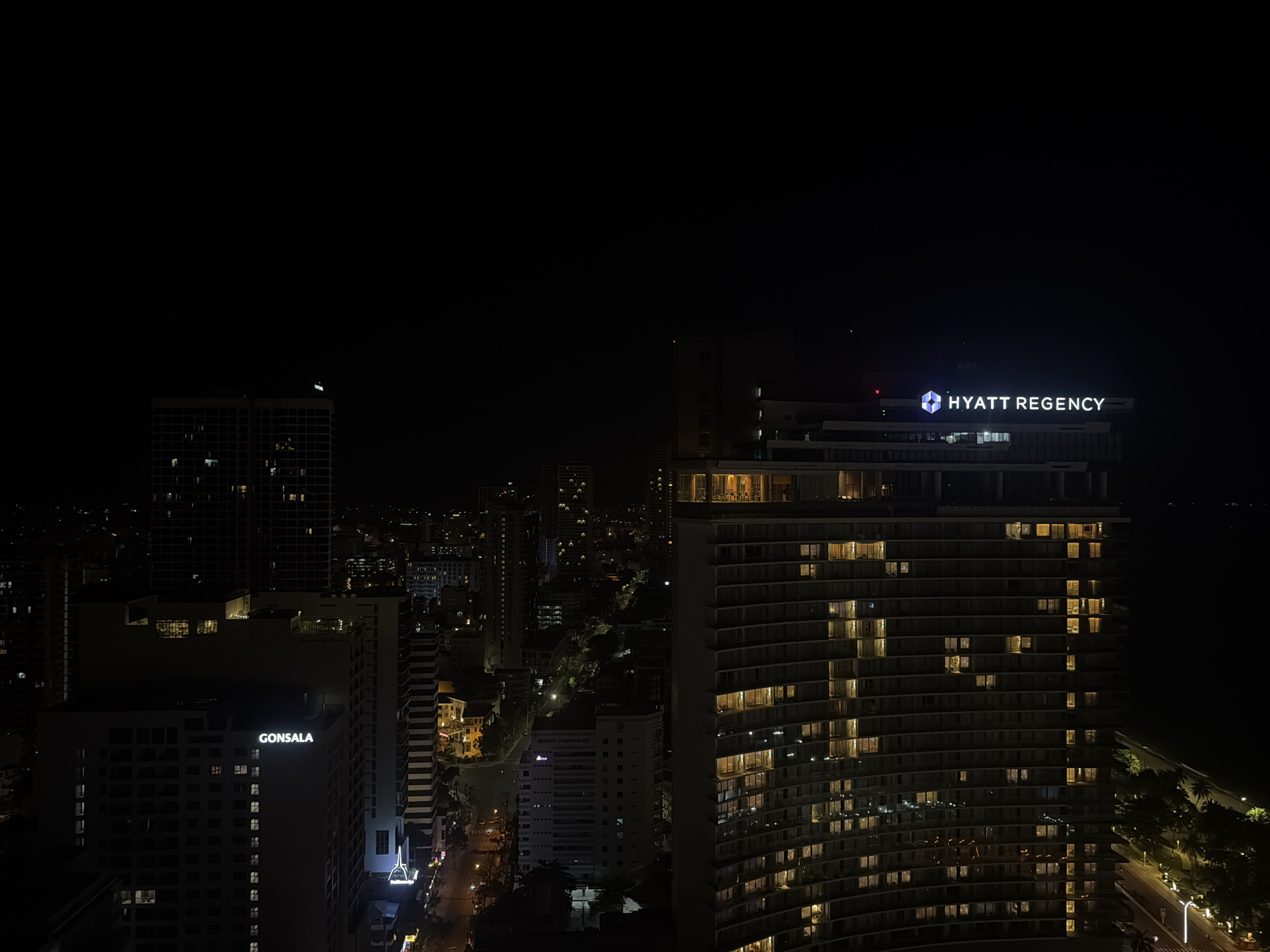 Night view of the city of Nha Trang Ward, Vietnam. Illuminated buildings dominate the scene. The Hyatt Regency hotel is prominently displayed, with lighted windows. Another building, with the sign "GONSALA", is also visible. Streets with lights meander through the city. Other buildings in the background are dimly lit. The sky is dark.