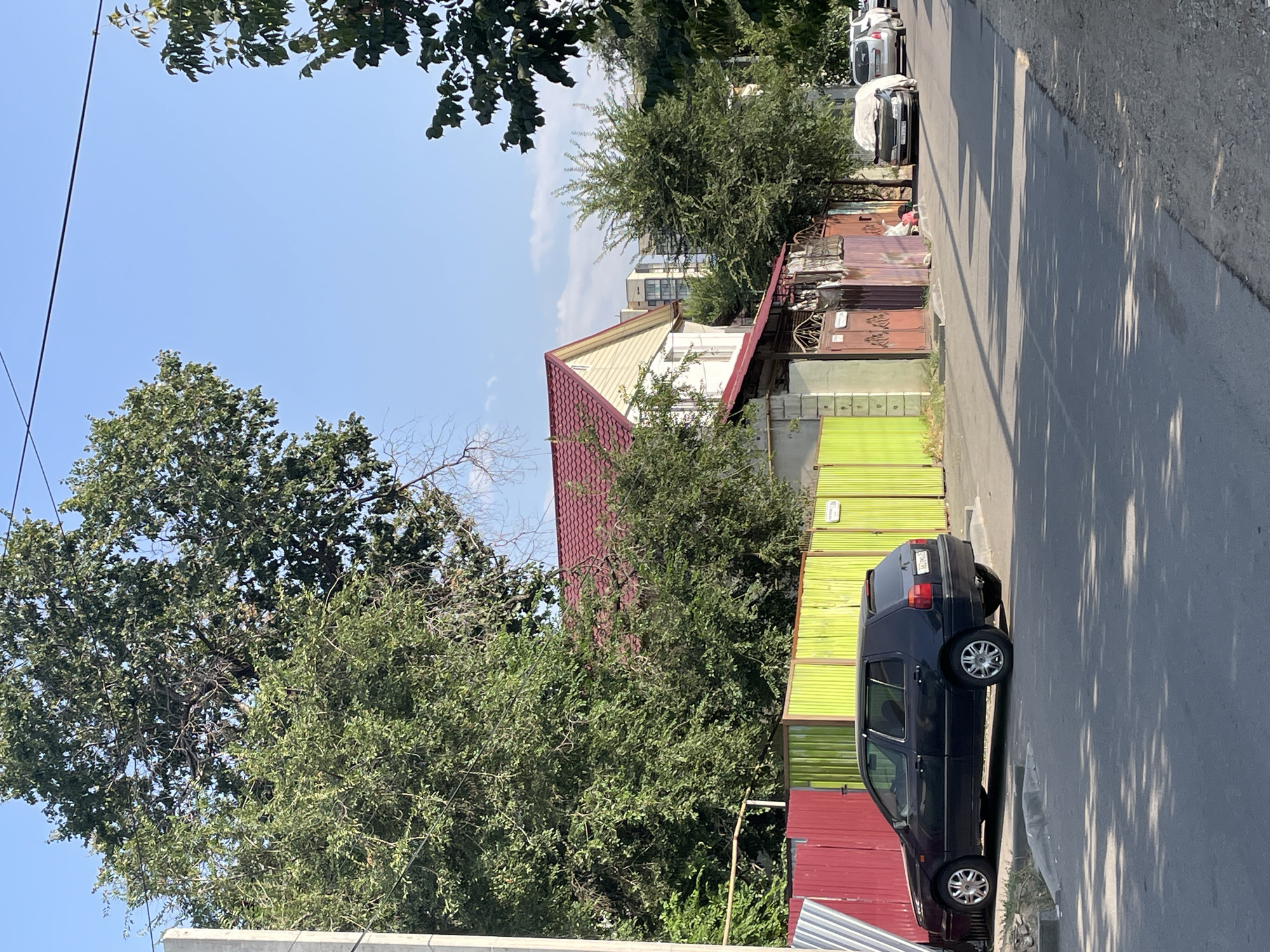 A dark blue hatchback is parked on the right side of a paved road in a residential area. The car is angled towards the curb, with its rear facing away from the camera. A multi-colored fence, featuring sections of red and bright green, runs along the property behind the car. A two-story house with a red-tiled roof is visible behind the fence. In the background, several modern apartment buildings stand tall against a clear blue sky. The scene appears to be in Almaty, Kazakhstan, based on the car's license plate, which reads "682 ZHG 05." The sunlight casts shadows across the road, indicating it is daytime.