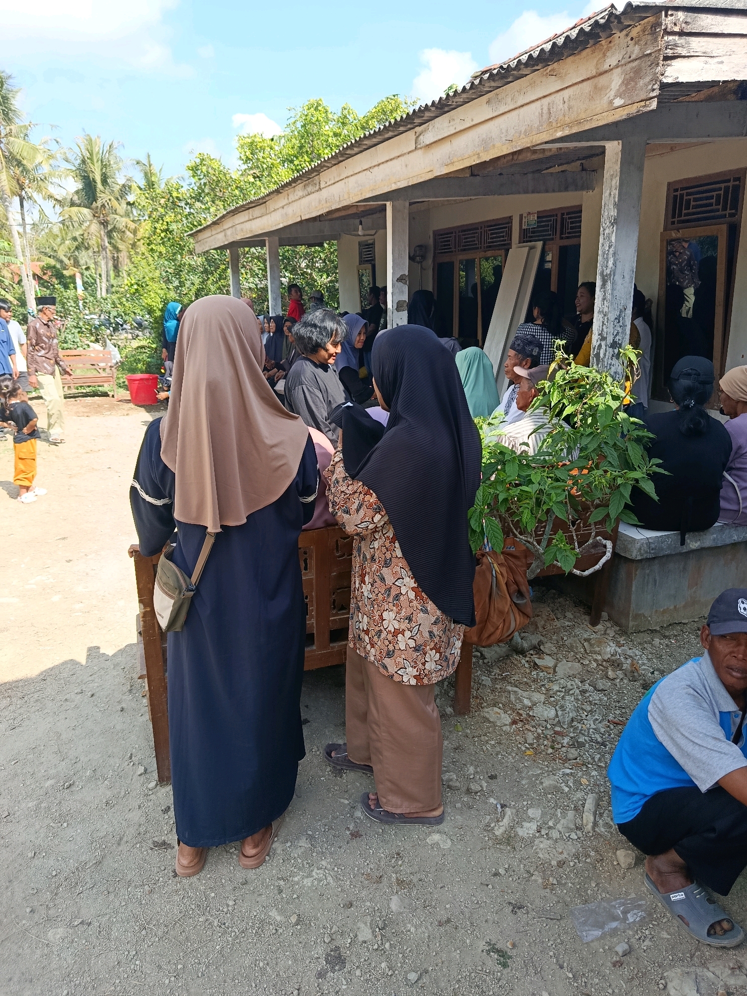 The image captures a daytime outdoor scene in Desa Bringkeng, Indonesia. In the foreground, two women wearing hijabs are standing. The woman on the left wears a brown hijab and a dark blue dress with a light-colored bag slung across her body. The woman on the right wears a black hijab and a patterned top with brown pants. Behind them, a group of people are gathered under the awning of a building with a corrugated metal roof. The building appears to be a house or a community gathering space. To the left, a man in a brown shirt and light-colored pants walks past a wooden bench, and a young child in an orange outfit stands near him. The environment is rural, with palm trees and lush green vegetation visible in the background. The ground is covered in dirt and small stones. A man in a blue shirt and black pants is squatting in the lower right corner of the frame, looking towards the camera.
