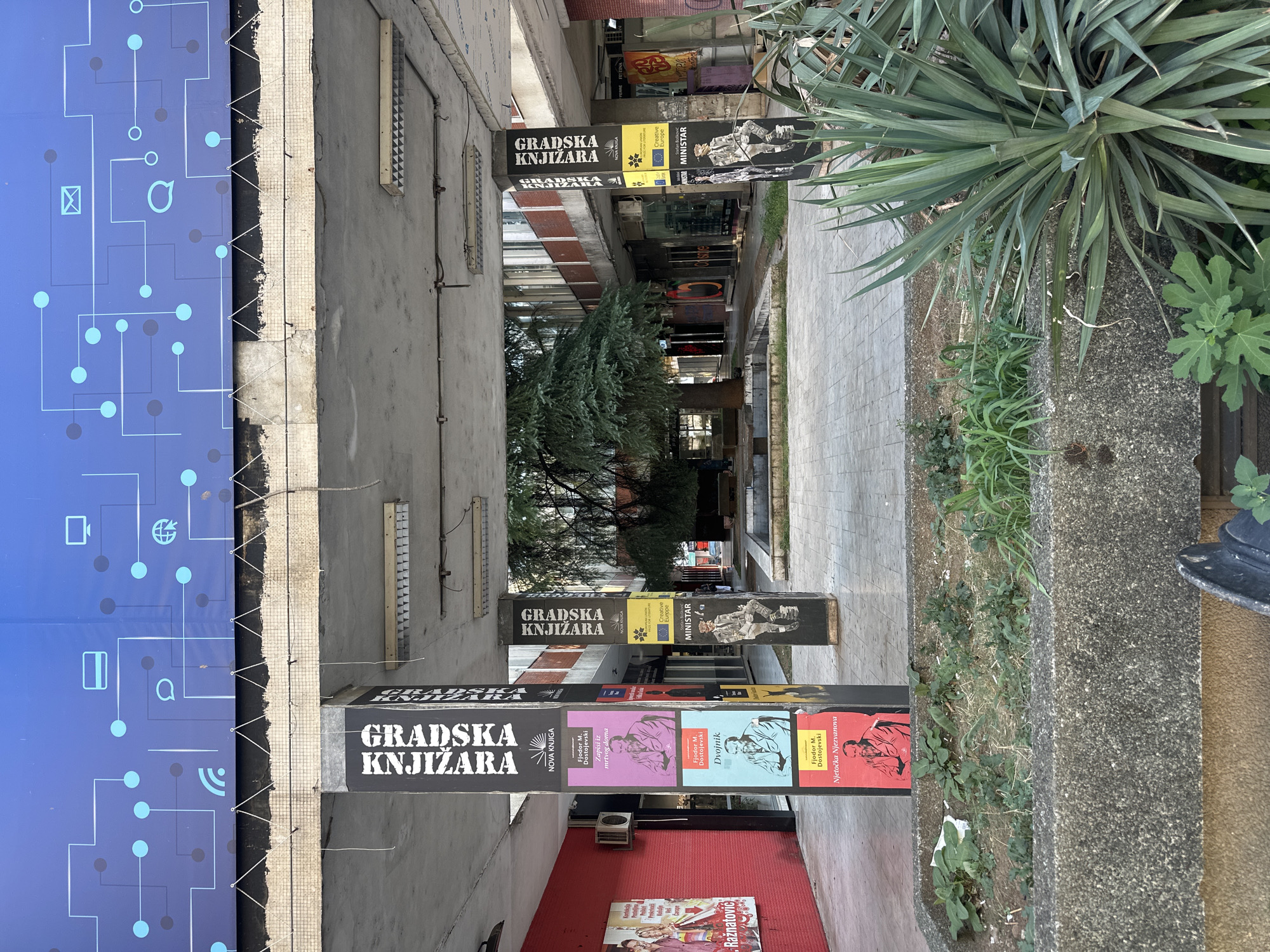The image shows an outdoor walkway with shops on either side. In the foreground, there are several concrete planters filled with vegetation, including a large yucca plant. The walkway is paved with light-colored tiles. On the left side of the walkway, there is a red-painted building with posters displayed on its exterior. In the center of the walkway, there are four large, black pillars with posters attached to them. These pillars appear to be part of the building's structure, providing support for an overhead canopy. The posters on the pillars advertise books and feature portraits of authors. Behind the pillars, there is a central courtyard with a large tree, a small fountain, and more shops. The overall impression is one of a somewhat neglected urban public space. This location is the courtyard of the "Gradska Knjizara" (City Bookstore) in Podgorica, Montenegro, recognizable by the repeated signage on the pillars. The overhead structure appears to be a canopy covering the walkway, and a large blue banner with a digital network motif is attached to the underside of the canopy. The lighting suggests it is daytime, with the sun casting shadows.
