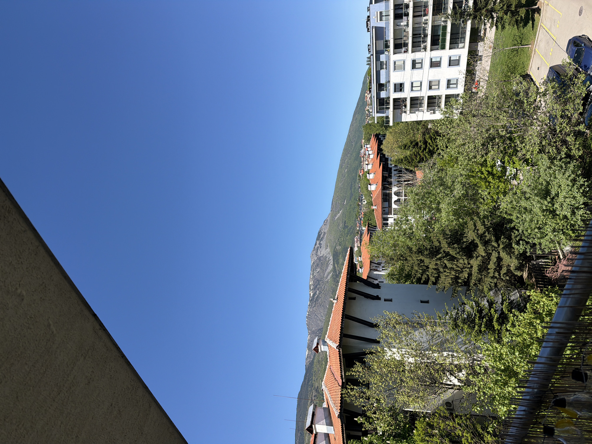 The image shows a view from an elevated position, possibly a balcony in Sofia, Bulgaria. A building with a red-tiled roof is visible, along with a modern white apartment building. In the background, a mountain range covered with green vegetation is visible under a clear blue sky. Green trees are also present, partially obscuring the view of the buildings. A wall or side of a building is visible on the left side of the frame. The sky occupies a significant portion of the image.