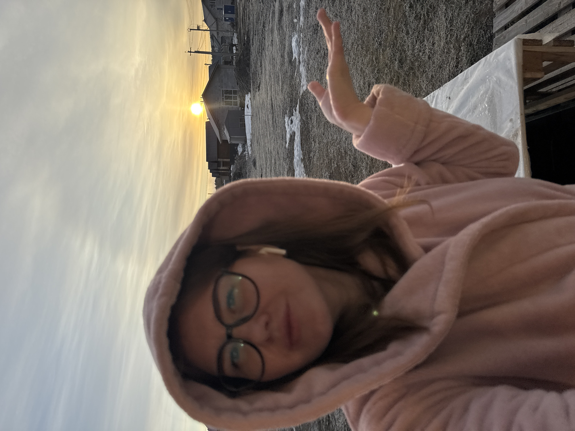 A selfie-style photograph shows a woman with glasses and a pink hooded robe. She is wearing a wireless earbud in her left ear and making a hand gesture. The background features a landscape including houses, utility poles, and a sky with a setting or rising sun. The ground has a patchy appearance, possibly snow or frost. The location is Nestiukovo, Russia, based on the prompt.