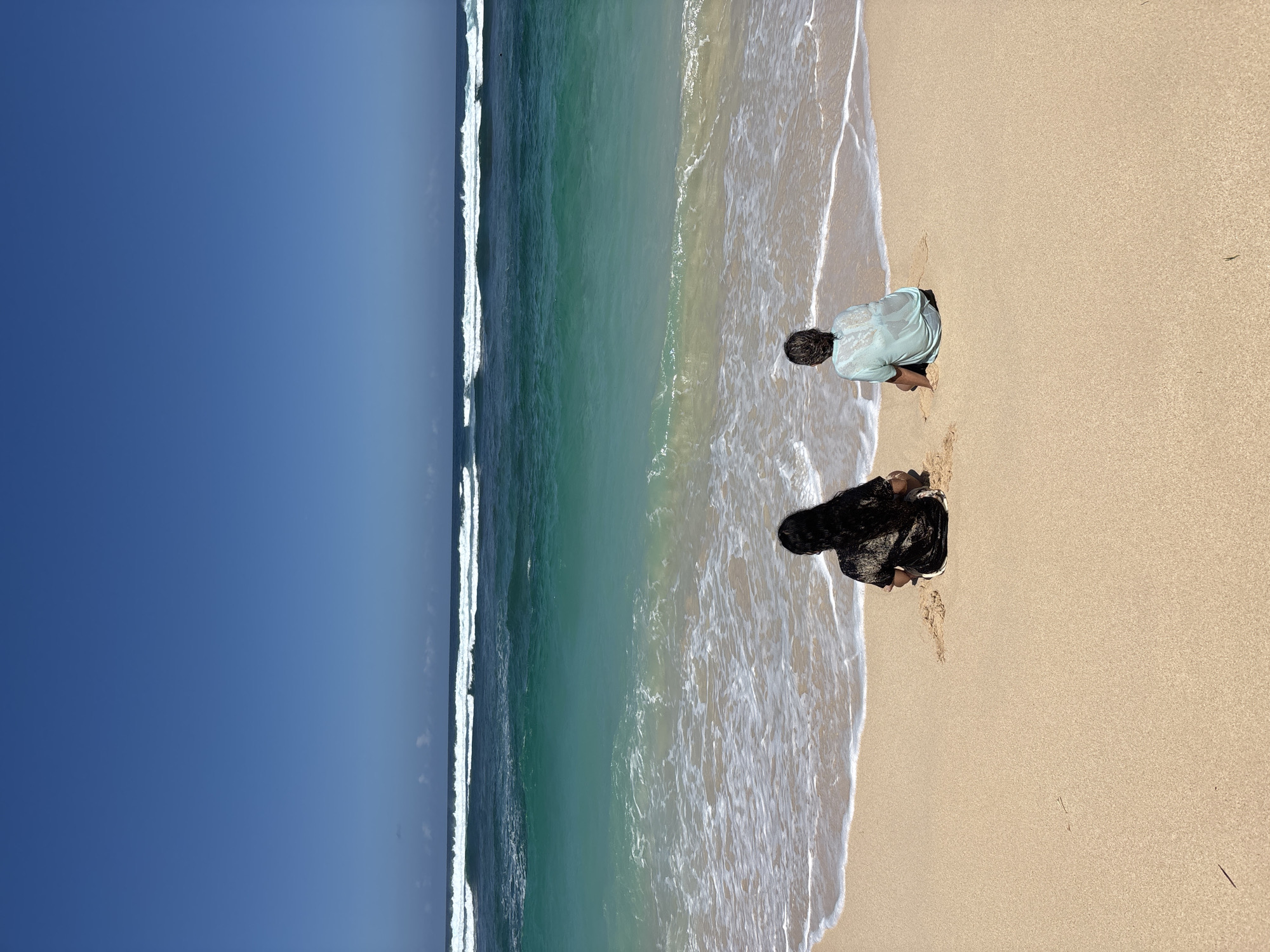 Two individuals are seated on a sandy beach facing the ocean in Kuta Selatan, Indonesia. The person on the left has long, dark hair and is wearing a dark, patterned shirt. The person on the right is wearing a light blue, wet t-shirt. Both individuals are sitting with their backs to the camera. The ocean is a vibrant turquoise color with white waves crashing gently on the shore. The sky is clear and blue. The sand is light brown.