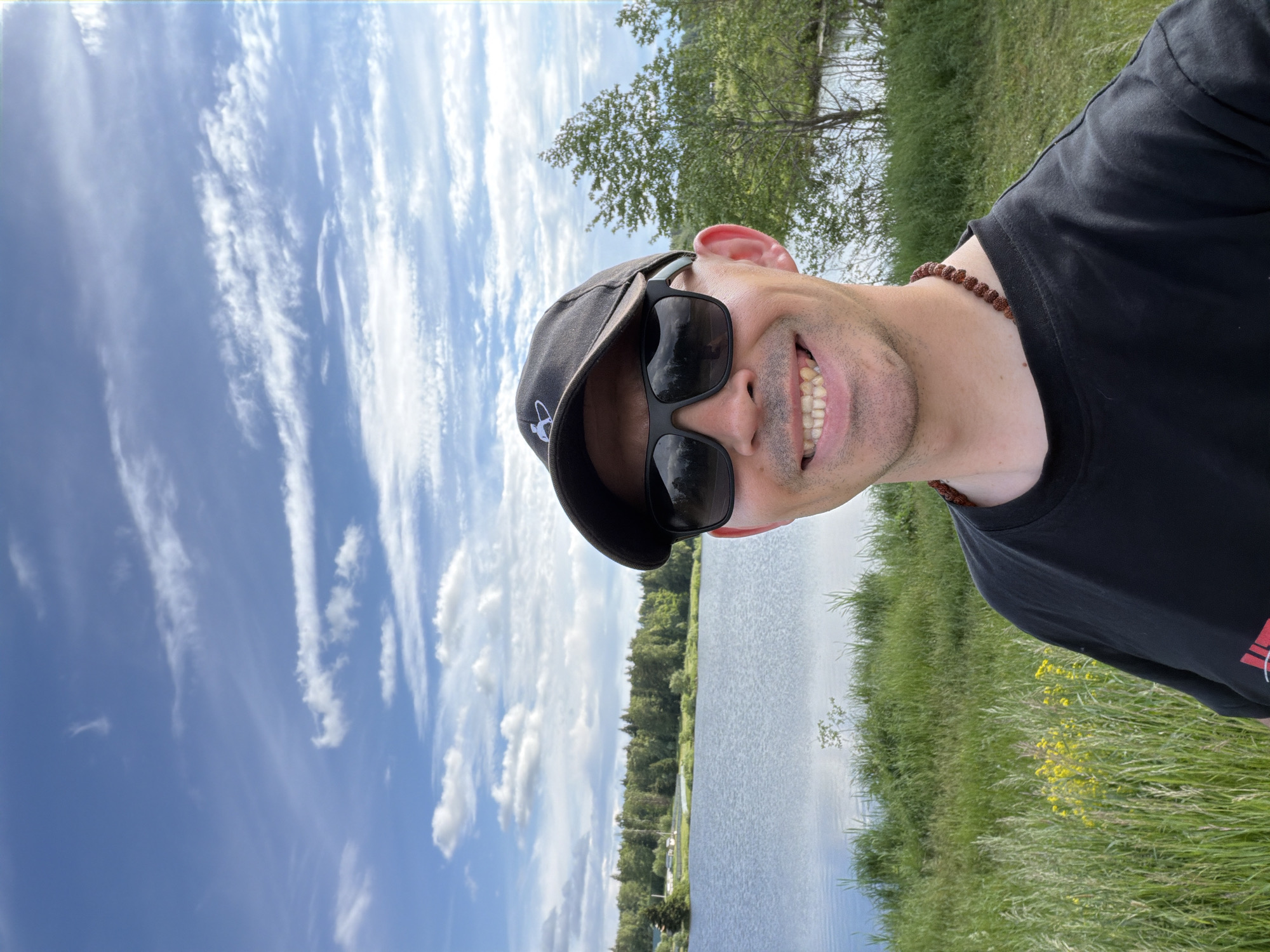 A selfie-style portrait captures a man smiling broadly while outdoors in Stolyarovo, Russia. He is looking directly at the camera, wearing a black baseball cap, dark sunglasses, and a black t-shirt. A brown beaded necklace is visible around his neck. His wide smile reveals his teeth, conveying a cheerful and relaxed emotion.
The background presents a serene natural landscape. A large body of water, likely a lake or river, spans the middle ground, reflecting the bright sky. The banks are covered in lush green grass and yellow wildflowers in the foreground, transitioning to a dense treeline of various deciduous and coniferous trees in the distance. Above, the sky is a vibrant blue with scattered white, wispy clouds, suggesting clear and pleasant daytime weather. The overall scene depicts a peaceful moment enjoying the natural environment. No legible text is visible on the man's attire or accessories, though a small, unclear emblem is on his cap.