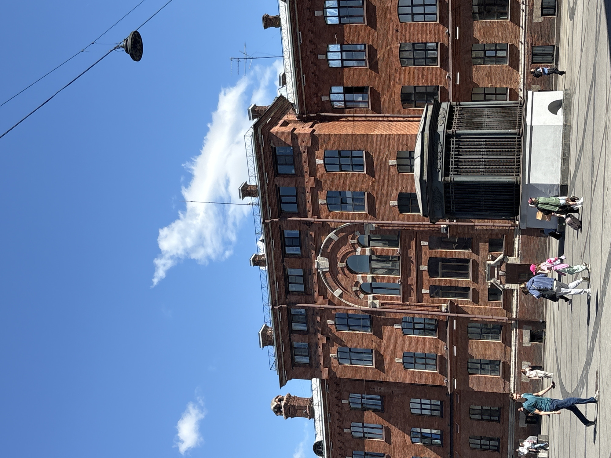 This is a vertical, eye-level medium shot of a brick building and a street scene, set in Saint Petersburg, Russia. The building is tall, made of reddish-brown brick, and features numerous dark-framed windows of various shapes and sizes. On the roof, there are chimneys and some antenna-like structures. A large, ornate metal gate or structure is visible near the bottom right of the building. A street lamp hangs from wires in the upper left of the frame.
Several people are visible on the paved street in front of the building. In the foreground, a man in a blue t-shirt and jeans is holding a phone and looking to his right. Further down the street, a group of three people, including a child in a pink hat, are walking. Another person is sitting on a white structure to the right of the gate. The sky is a vibrant blue with a few scattered white clouds, suggesting it is a clear, sunny day, likely late morning or early afternoon. The overall mood is casual and urban, with people enjoying a bright day outdoors. No visible text is present in the image.