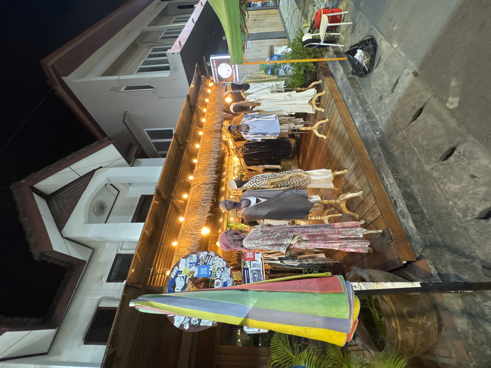 This image captures a street-level view of a boutique store at night in Koh Phangan, Thailand, specifically in the Tambon Koh Phangan. The store features a wooden platform display area showcasing mannequins dressed in bohemian-style clothing. The mannequins are arranged with a variety of garments, including flowing dresses, layered tops, and patterned attire. Above the display, a thatched roof is illuminated by strings of warm-toned lights, creating an inviting ambiance. A colorful, striped umbrella stands prominently on the left side of the frame. In the background, the facade of a multi-story building is visible, with windows and architectural details illuminated. To the right, a green canopy extends over a section of the street, and a white plastic chair and a red bucket are placed near the sidewalk. The street itself is paved with concrete, showing some wear and tear, including open grates. A black garbage bag sits on the sidewalk in front of the store.