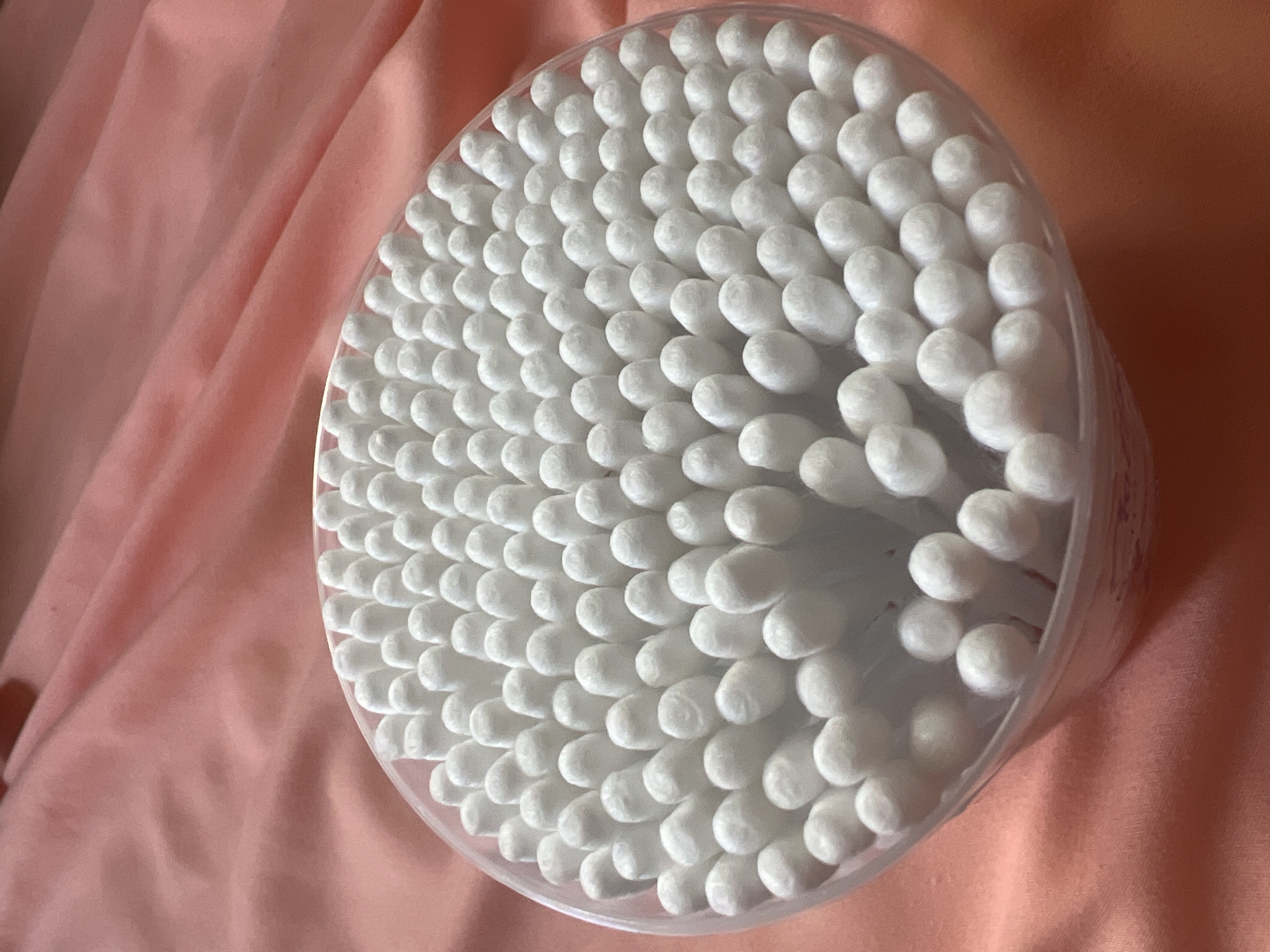 The image shows a clear plastic container filled with cotton swabs, arranged in a dense, somewhat circular pattern. The swabs are white, with rounded cotton tips. The container is placed on a wrinkled, pale pink fabric that appears to be silk or satin, casting soft shadows. The lighting is soft and diffused, highlighting the texture of both the cotton swabs and the fabric. The background is entirely composed of this pink fabric, with some areas appearing darker due to folds and shadows. There are no people or animals visible in the image. The image does not provide any information or context that would allow for its location to be determined in the city of Pskov, Russia.