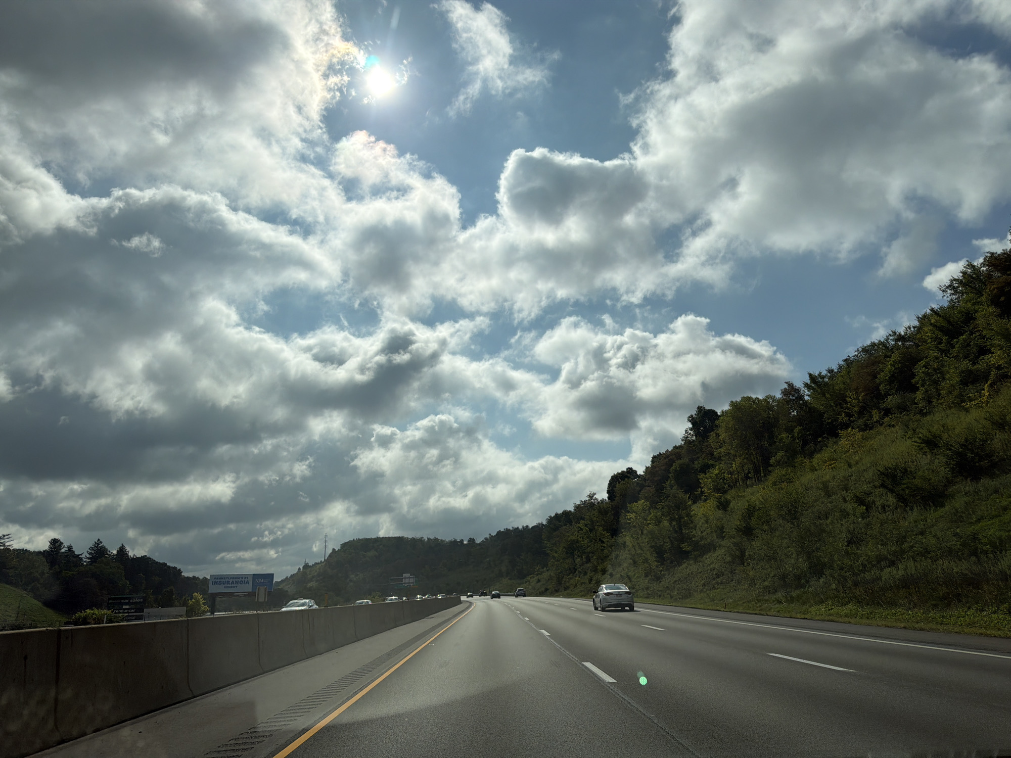 The image depicts a view from a vehicle traveling on a multi-lane highway, likely in Collier Township, United States of America. The highway is paved and features yellow and white lane markings. On the left side of the highway, a concrete barrier separates the lanes from a grassy embankment and a billboard. On the right side, a densely wooded hillside rises steeply. The sky is partly cloudy with bright sunlight visible through breaks in the clouds. Several vehicles are visible on the highway, including cars and trucks, all traveling in the same direction as the viewer's vehicle. The overall impression is of a typical highway scene on a day with variable weather conditions.