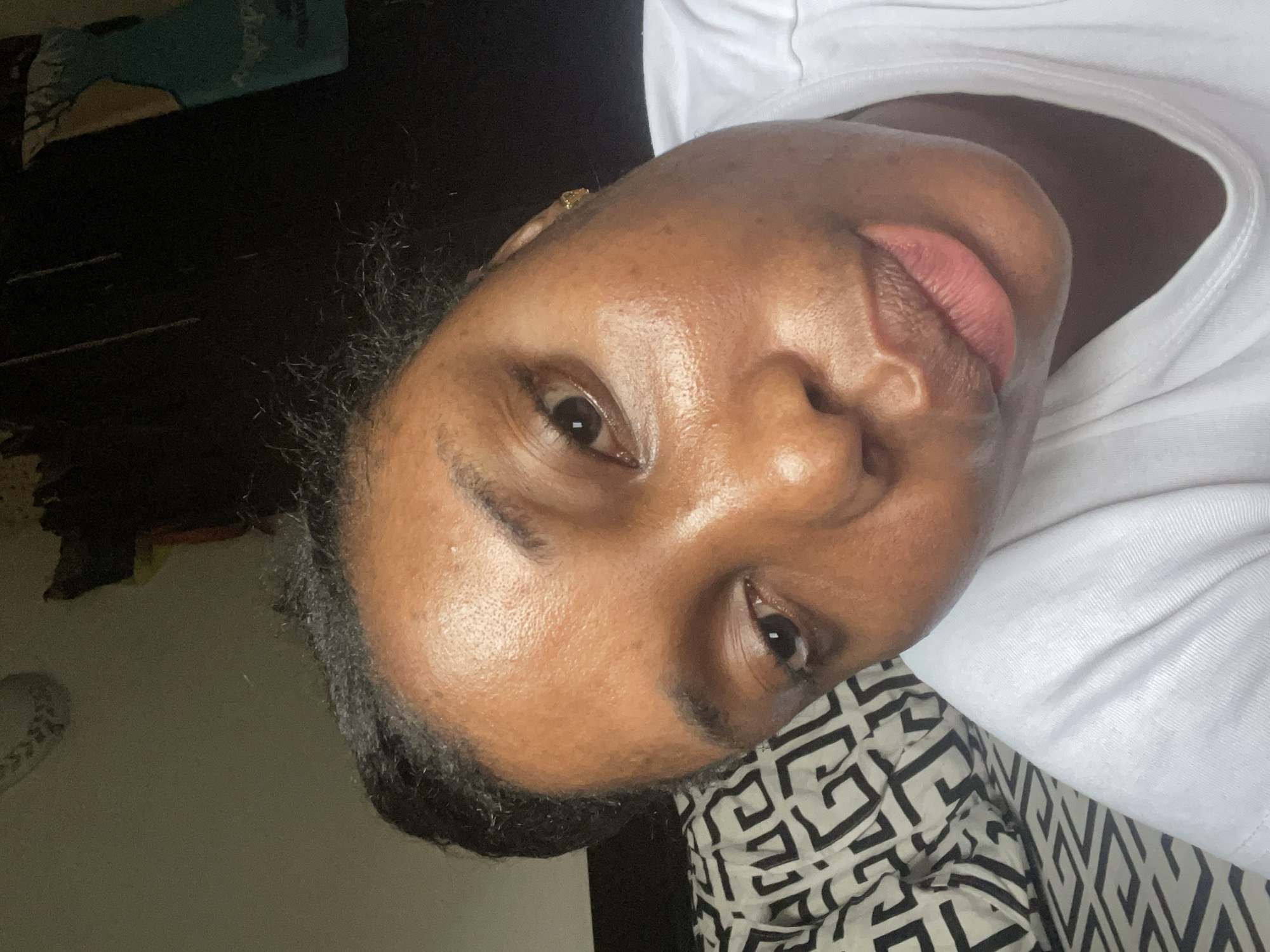 An eye-level portrait of a dark-skinned person with a white t-shirt. The person has short, dark hair, brown eyes, and pinkish lips. A gold earring is visible on the left ear. The background includes a black surface and a patterned black and white fabric. The image does not contain enough information to verify the location of the city of Abuja, Nigeria.
