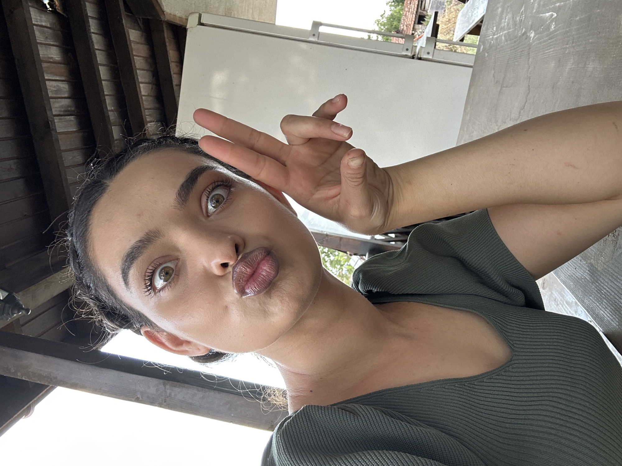 A young woman with dark hair styled in two buns, light brown eyes, and full lips pursed for a kiss, is pictured in a close-up selfie. She is wearing a green, ribbed, V-neck top. Her right hand is raised, with two fingers extended in a peace sign. Her left arm rests on a wooden table. The background suggests an outdoor setting, possibly a porch or patio, with a wooden ceiling structure above and a white refrigerator to the right. Hints of greenery and a rustic building are visible in the distance. The overall ambiance is casual and informal. The image was captured in Malevo, Bulgaria.