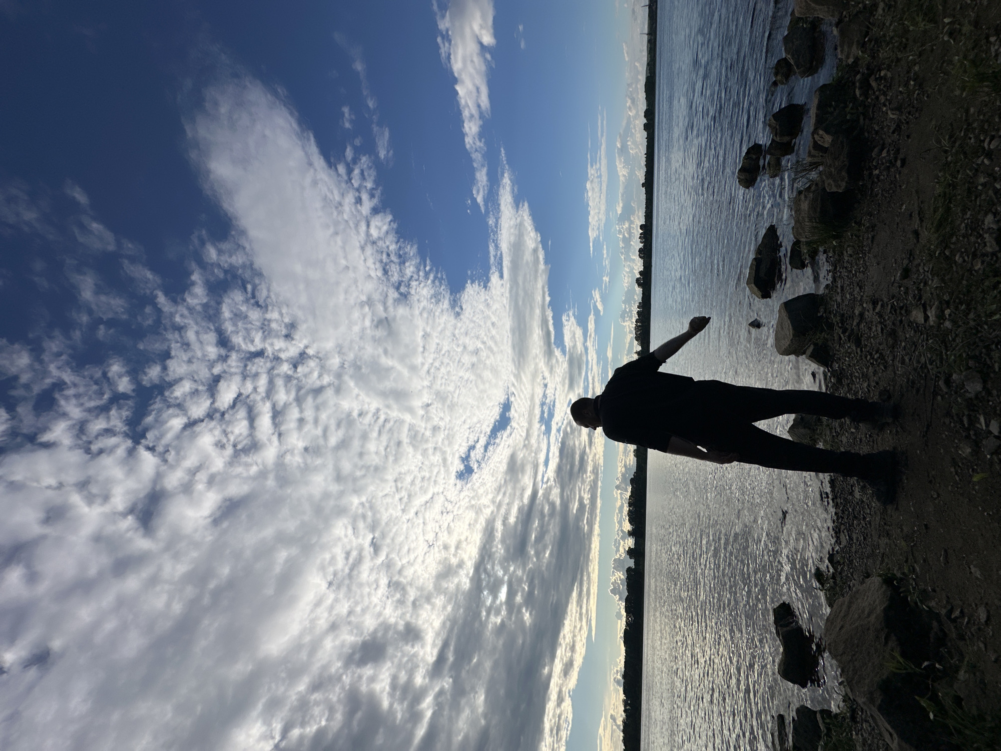 A silhouette of a person stands on the rocky shore of a wide river, facing away from the camera and towards the water. The person is wearing a dark, possibly black, t-shirt and pants, and appears to be holding a small object in their right hand, which is extended towards the water. The river surface is rippled, reflecting the dramatic sky above, which is filled with a mix of dark, textured clouds and patches of bright blue sky with scattered white clouds. The opposite bank of the river is lined with a dark, silhouetted treeline, suggesting a natural landscape. The foreground is composed of dark soil and rocks of various sizes. The scene appears to be set in the late afternoon or early evening, judging by the lighting. The location is the bank of the Kama River in the city of Chaikovsky, Russia.