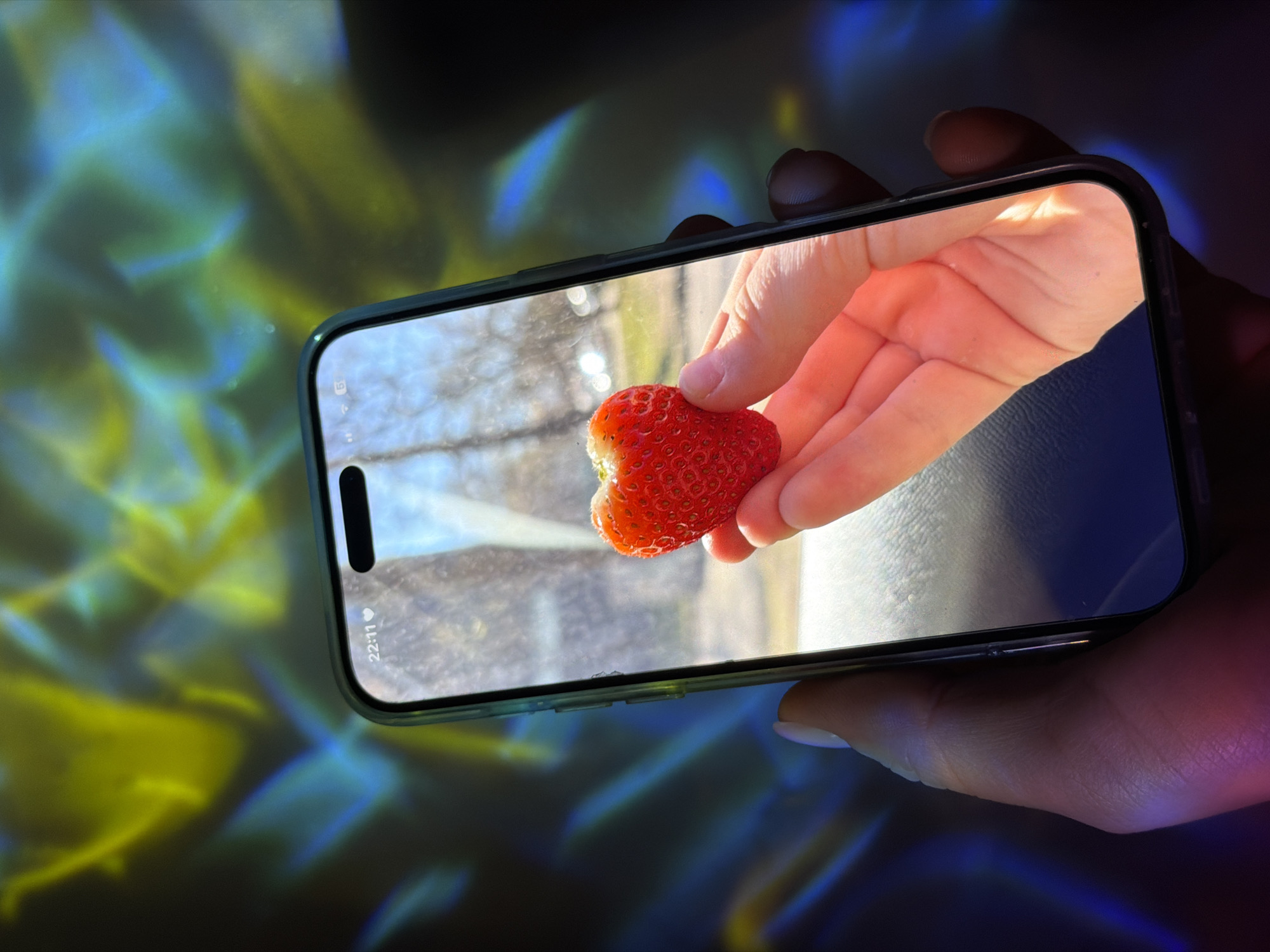 A hand holds a smartphone displaying a picture of a hand holding a heart-shaped strawberry. The smartphone's screen displays a bright, daytime scene with a blurry background suggestive of trees. The time on the phone is 22:11. The background of the image outside the phone screen consists of blurred streaks of blue and yellow light. The setting and location in Dreiliņi, Latvia, cannot be definitively ascertained from this image.