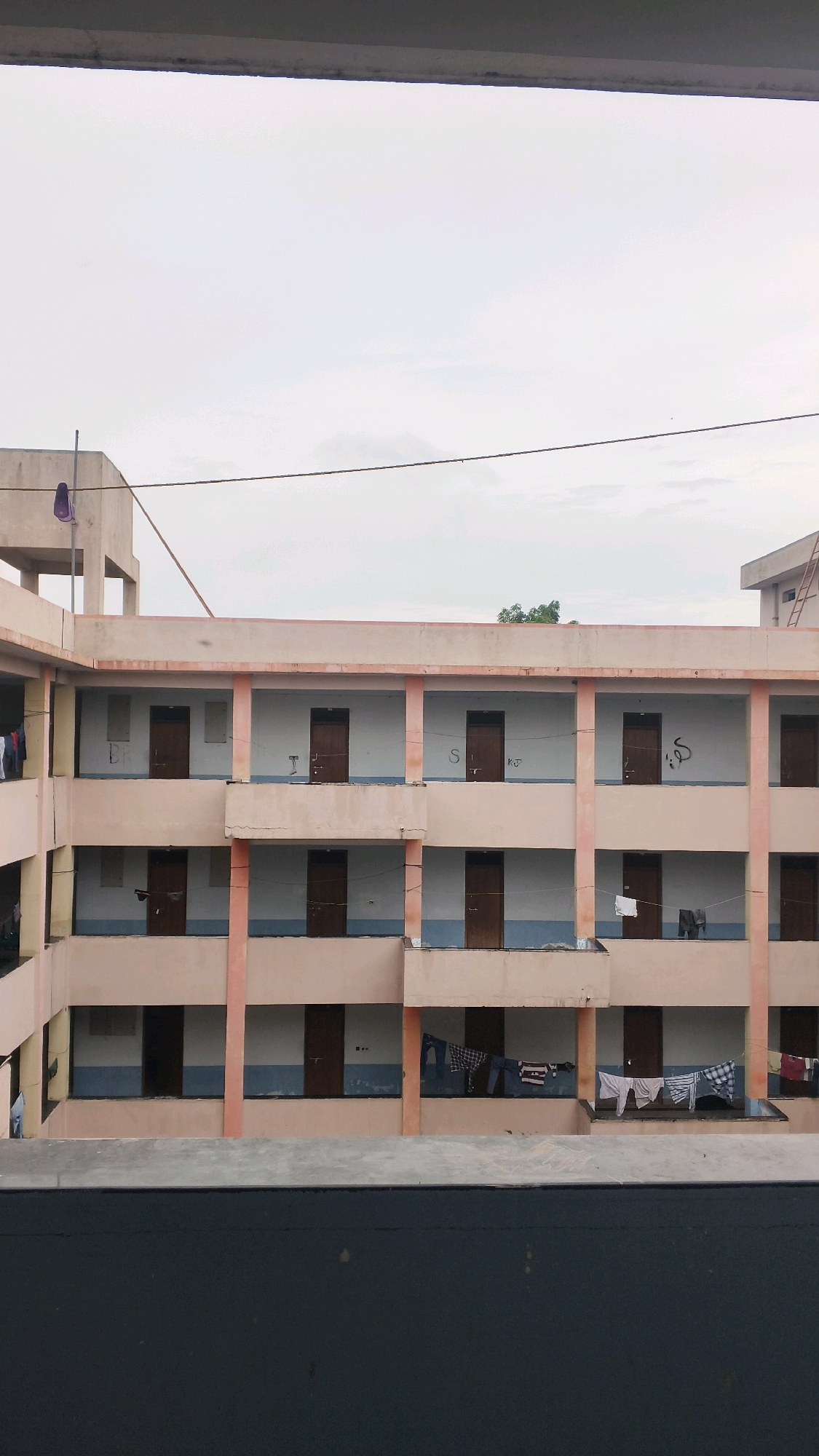 The image depicts a multi-story building, likely a dormitory or hostel, with rows of identical rooms visible. The exterior is painted in muted tones of peach and blue. Each room appears to have a wooden door and a window or opening onto a shared balcony or corridor. Laundry is hanging on clotheslines in front of some of the rooms on the lower floors. The sky is overcast, and there is a suggestion of trees and a ladder on the far right. The foreground shows a dark, textured surface, possibly a ledge or railing, obscuring the very bottom of the building. The overall impression is one of a functional, residential building in a somewhat overcast environment. The location is identified as Sarafpalle, India.