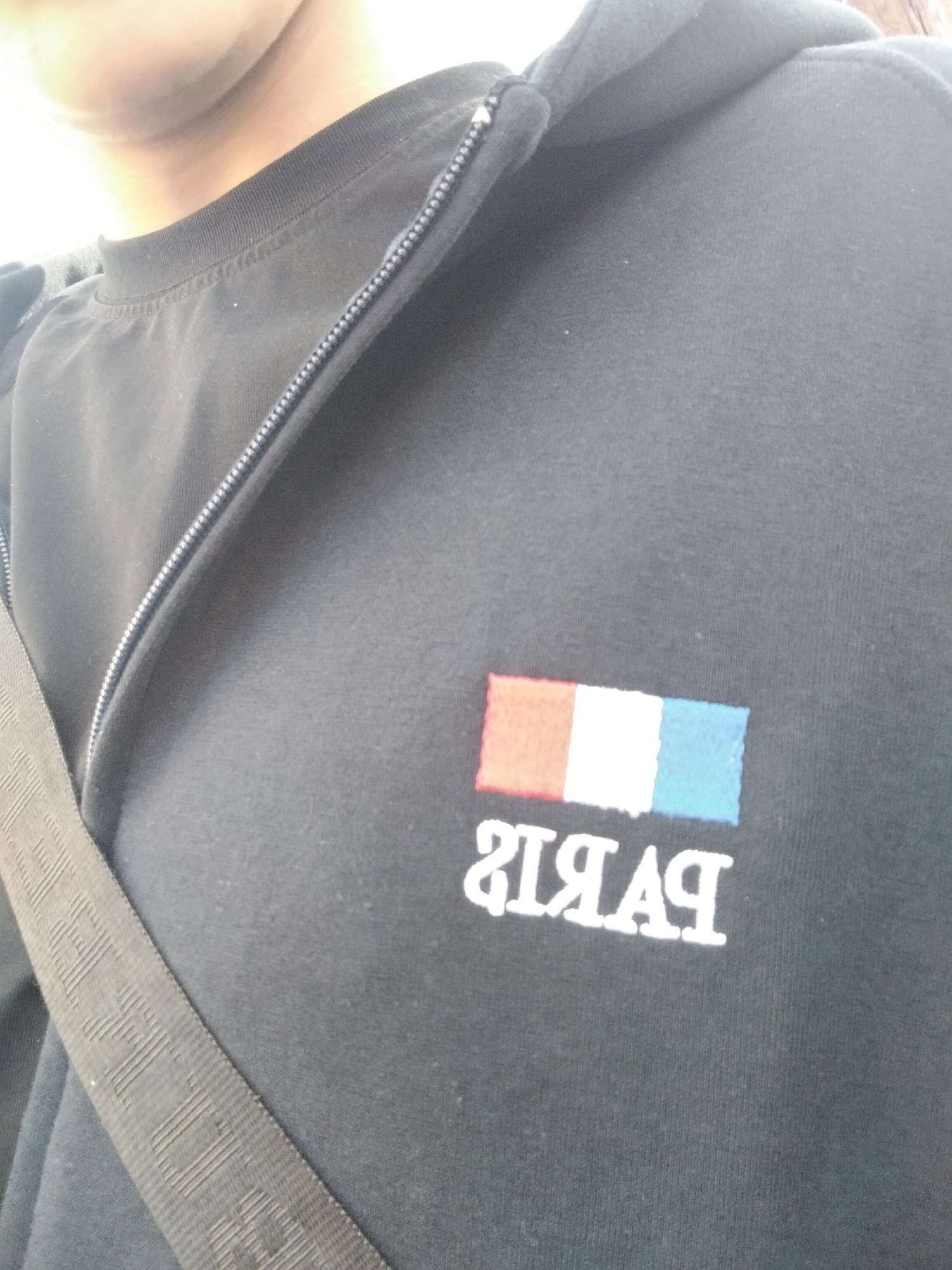 A person is wearing a black zip-up hooded sweatshirt with a French flag and the word "Paris" printed on the chest. The image is a close-up, capturing the upper part of the torso and part of the neck. A black strap is visible across the chest. The background is blurred. There is no evidence in the image to determine the location in Yerevan, Armenia.