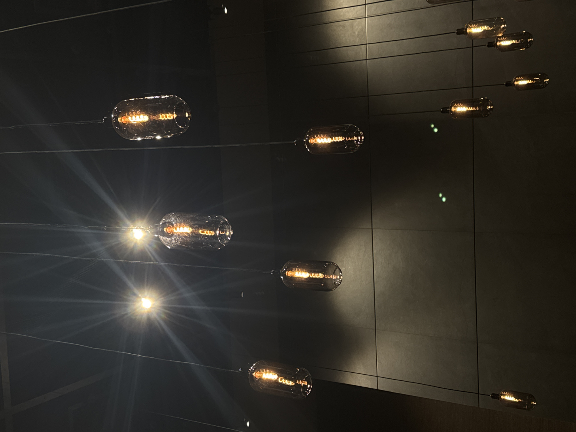 A downward-facing medium shot captures a cluster of vintage-style incandescent light bulbs hanging from thin black wires against a dark, textured wall. The bulbs, enclosed in clear glass casings, have visible filaments emitting a warm, orange glow. Some bulbs are brighter, with light rays emanating outwards, suggesting they are closer to the camera or more intensely lit. The wall behind them appears to be made of large, rectangular panels with a subtle sheen. The lighting suggests an indoor setting, possibly a restaurant or modern interior design space. There are no people visible in the image. The overall mood is one of sophisticated, ambient lighting.