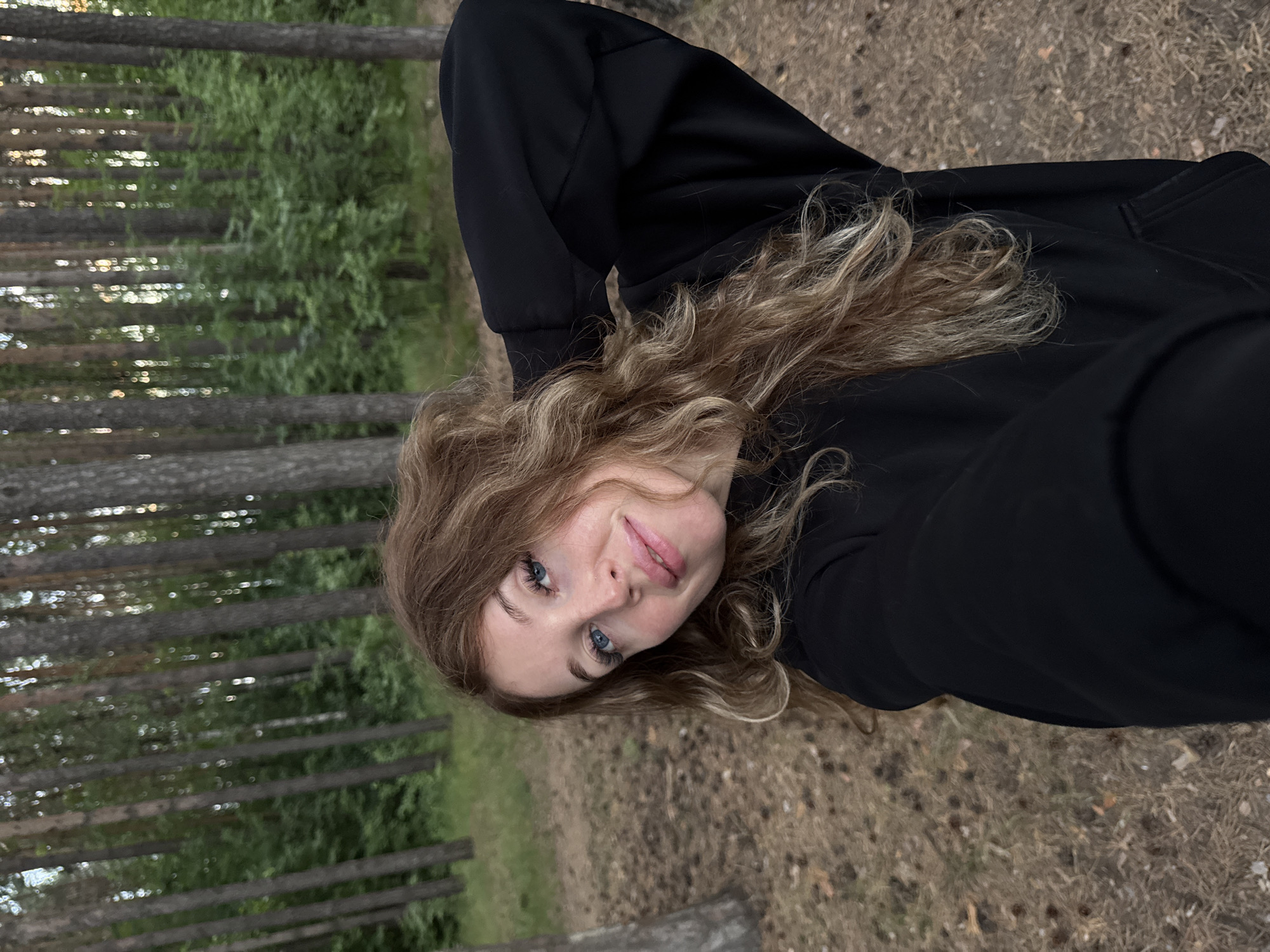 A young woman with long, wavy auburn hair and blue eyes is taking a selfie in a forest. She is wearing a black hoodie and has her right arm behind her head. The background is a dense forest with tall, thin trees and green foliage. The ground is covered with pine needles and fallen branches. The lighting suggests it is either early morning or late afternoon. The location is the coniferous forest on the outskirts of Chaikovsky, Russia.