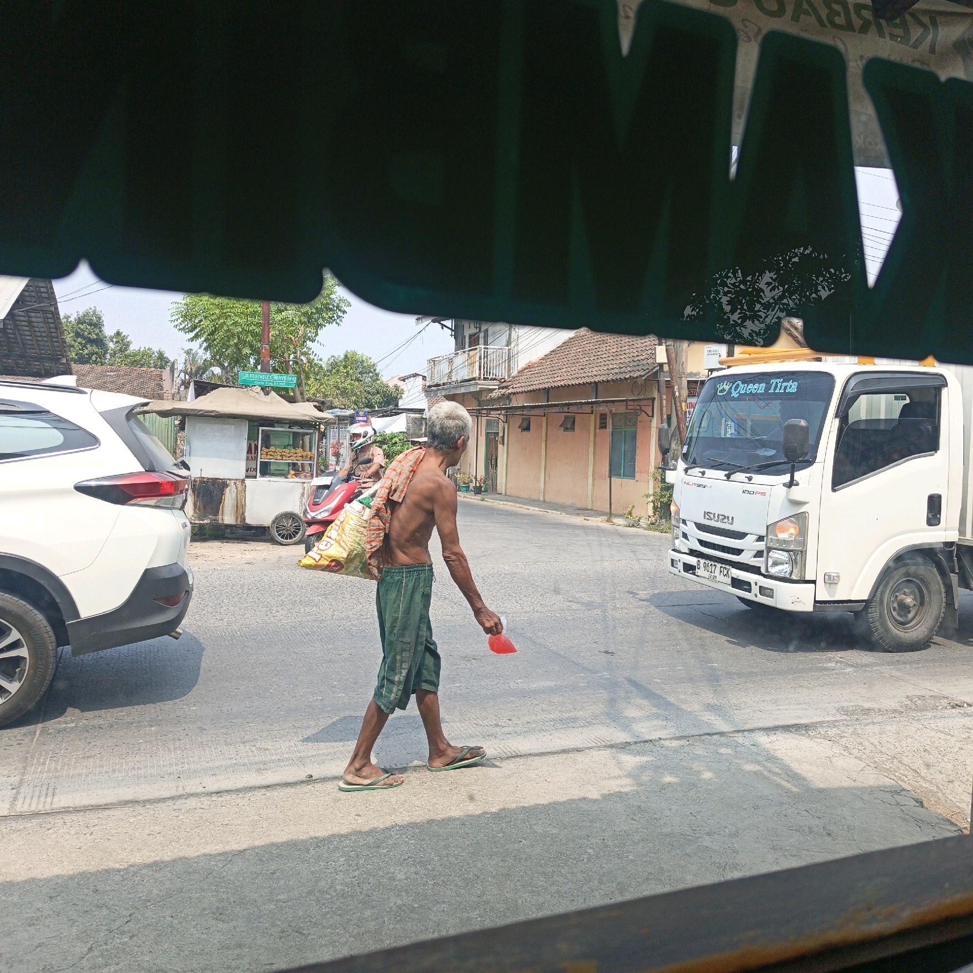A man walks across a street in Pegandon, Indonesia, carrying a large, yellow woven bag over his right shoulder and a red plastic bag in his right hand. He is shirtless, wearing green shorts, and flip-flops. He is walking from left to right in the frame, towards the middle of the street. In the background, a white Isuzu truck is driving away from the camera. To the left of the truck is a white food stall with a brown roof. A red motorcycle is parked in front of the food stall, with a person wearing a helmet sitting on it. Behind the food stall and motorcycle are buildings and trees. The scene is illuminated by daylight.