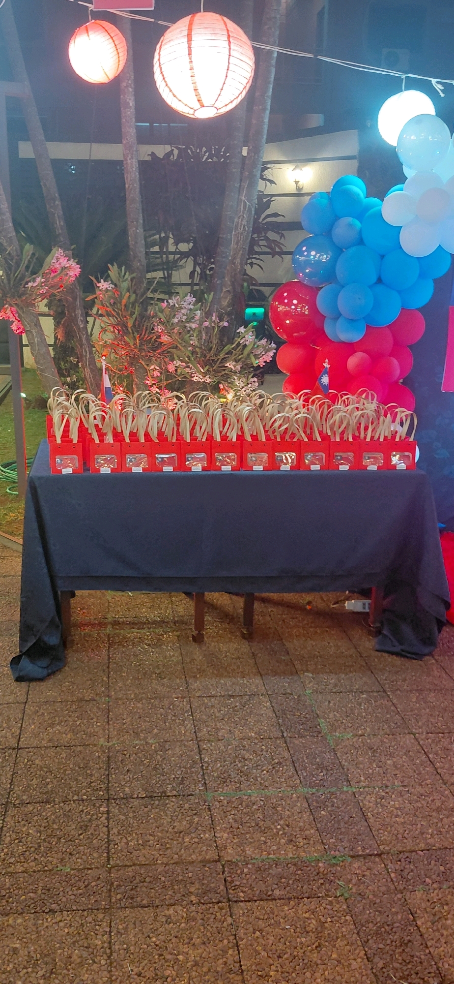 The image shows a table covered with a dark blue cloth, set up outdoors. On the table, there are numerous red gift bags with handles made of natural fiber. Each bag has a clear window on the front, revealing small items inside, possibly toys or favors. A small flag, appearing to be the flag of Taiwan, is placed in front of one of the gift bags. Behind the table, there are trees with some flowering plants. In the background, there are several round paper lanterns, two orange and one white, hanging from strings. To the right, there is a cluster of balloons, predominantly blue with some red and white ones. The ground is paved with small, reddish-brown stones. The overall scene suggests an outdoor celebration or event. The location is identified as Encarnacion, Paraguay, implying this is likely a festive gathering in that city. There are no people visible in the frame.