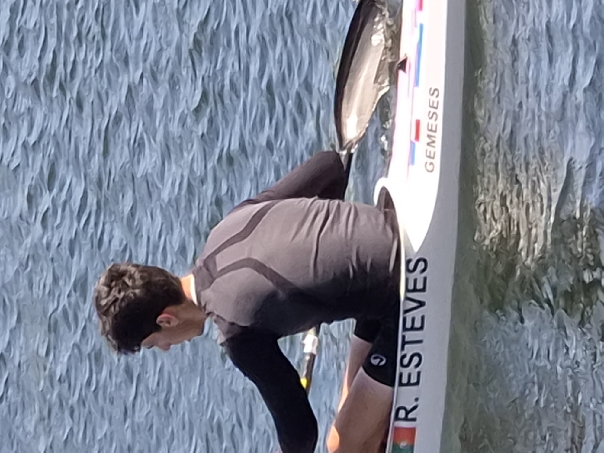 A man is rowing a kayak on a body of water, likely the Cávado River in Esposende, Portugal, as the name "ESTEVES" and the Portuguese flag are visible on the side of the kayak. The kayak also has the word "GEMESES" on it. He is wearing a long-sleeved grey top and black shorts. He is in a low stance within the kayak, holding a paddle with both hands. The water has a rippled texture.