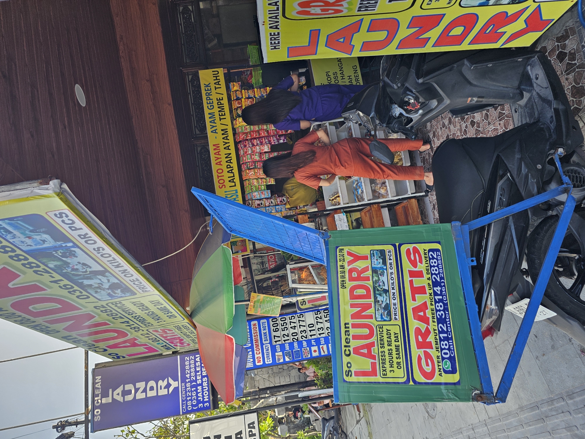 Here's a detailed description of the image:
**Location:** The scene appears to be on a street in Denpasar, Indonesia, based on the presence of Indonesian script, currency exchange rates, and the general street scene.
**Subjects:**
* Two women are bent over, looking at items displayed in front of a small storefront. One woman wears a purple outfit and the other an orange outfit.
* Motorbikes are parked on the street.
**Objects:**
* "So Clean LAUNDRY" signs in various forms, including vertical signs, sandwich boards, and promotional material.
* A money changer sign displaying currency exchange rates.
* A food stall selling "Soto Ayam - Ayam Geprek," a traditional Indonesian dish.
* Merchandise on display in the food stall and possibly the laundry business.
* A green and red umbrella.
**Actions/Interactions:**
* The two women are browsing or shopping at the storefront.
* The laundry service is promoted through signage.
* Motorbikes are parked, suggesting people have arrived to either shop or use the services.
**Environment:**
* The image is taken on a street with various businesses, including the laundry service and food stall.
* The environment appears to be a typical commercial area with businesses and street activity.
* A sign for laundry service is visible.