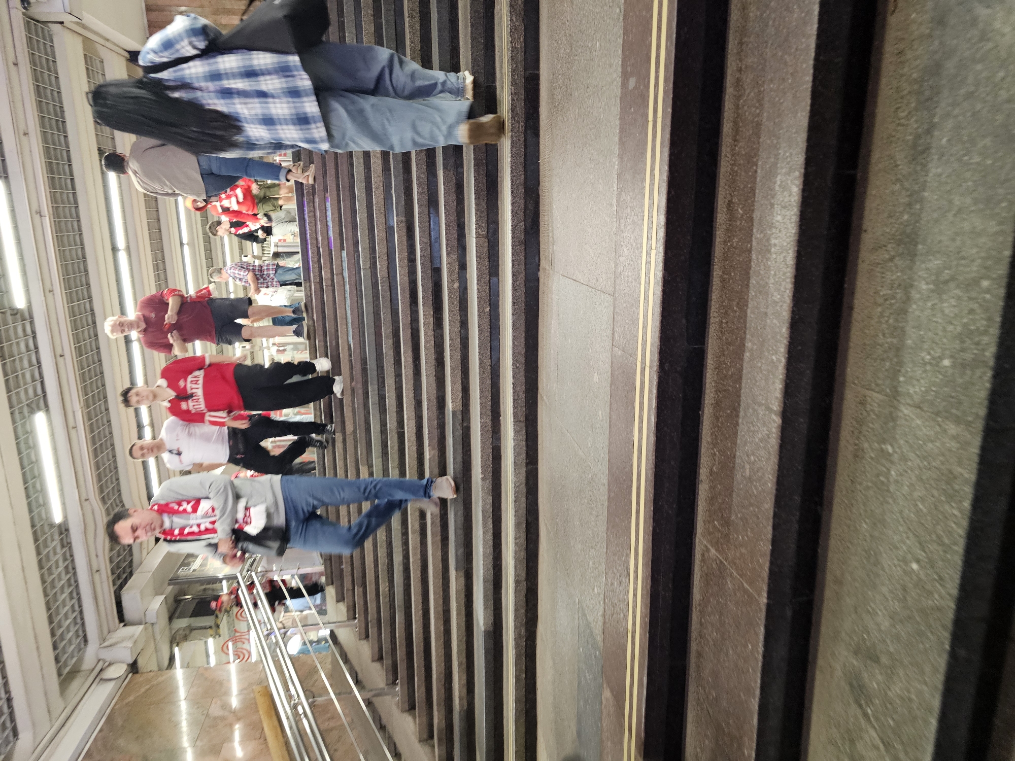 In a subway underpass in Khimki, Russia, a group of people are ascending a wide, multi-tiered staircase. The architecture of the underpass features a metallic ceiling with fluorescent lighting, and the walls are lined with polished stone. The staircase itself is constructed from granite-like material with yellow warning strips on some of the steps.
In the foreground, a woman wearing a blue and white plaid shirt and light blue jeans walks up the stairs, carrying a black bag over her right shoulder. Behind her, a man in a gray sweatshirt and blue jeans, adorned with a red and white scarf, is also ascending the stairs, holding a black bag. Further up the staircase, several other individuals are visible, including a man in a red t-shirt and black shorts, another in a white t-shirt and black pants, and others in casual attire, some wearing red clothing, potentially indicating support for a sports team. The overall scene suggests a public transit environment with a flow of pedestrian traffic.