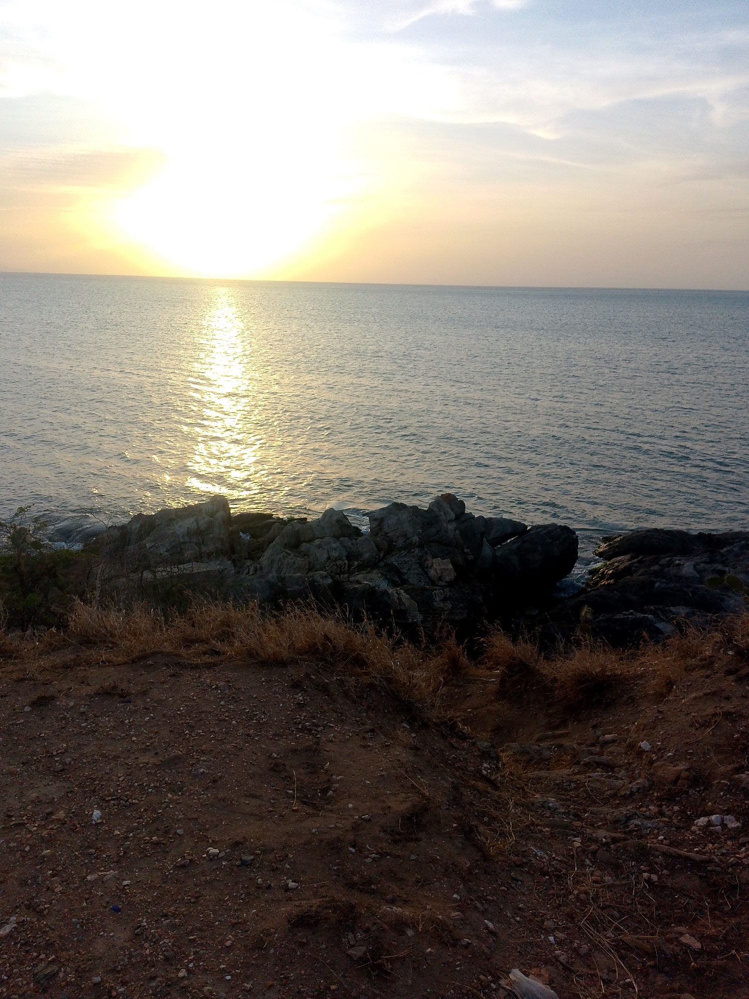 The image captures a sunset scene in Juan Griego, Venezuela, overlooking the ocean from a rocky promontory. The sun, a bright orb, is positioned near the horizon, casting a shimmering reflection on the water's surface. The sky exhibits a gradation of color, from bright near the sun to muted blues and whites further away. The foreground consists of a dirt area with sparse vegetation leading to a rocky shoreline. The rocks are dark and appear rugged. The ground is covered in dirt, small rocks and dry vegetation.