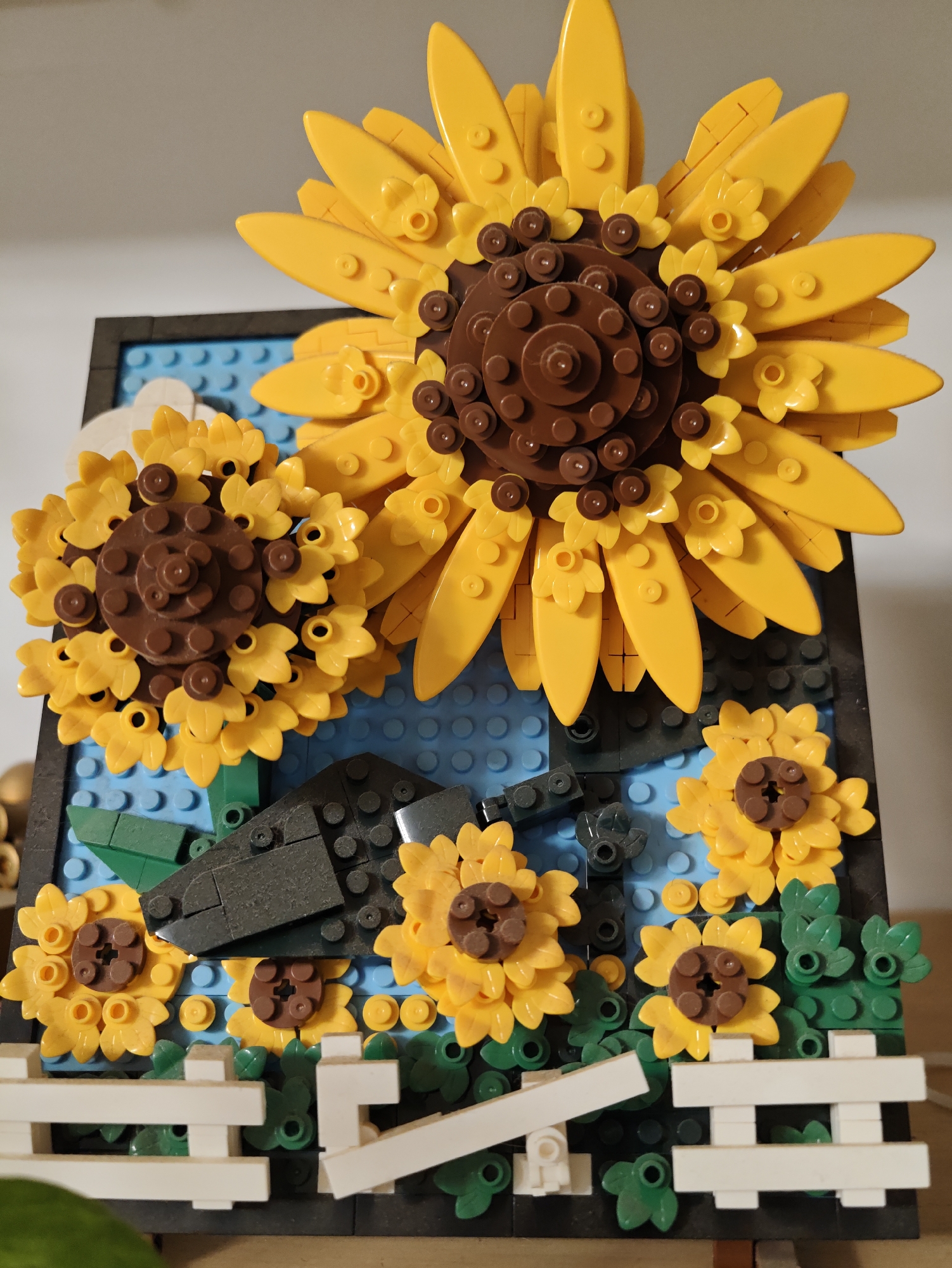 The image displays a close-up view of a LEGO diorama depicting a sunflower garden. The diorama features several sunflowers in various sizes, constructed from yellow and brown LEGO bricks. Smaller yellow flowers and green leaves are scattered throughout the scene. In the foreground, a white LEGO fence is visible. The background consists of blue LEGO bricks, possibly representing the sky. The diorama is set against a neutral, light-colored background. The overall composition suggests a handcrafted model, likely a display piece. The image does not contain any elements that would indicate a specific location in Petah Tikva, Israel, or any other city.