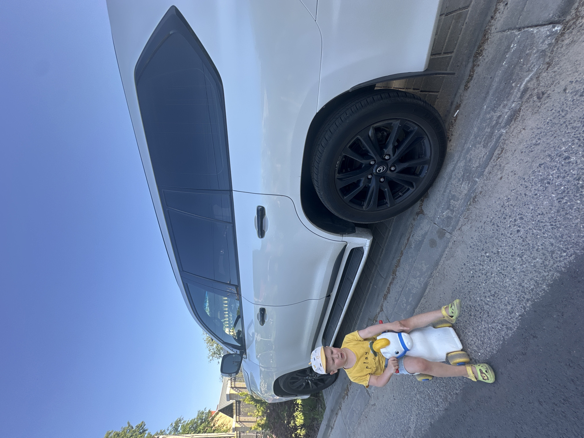 A young child is riding a white toy horse on a paved street next to a large, white SUV. The child is wearing a yellow t-shirt, shorts, a patterned baseball cap, and yellow sandals. The SUV is parked along the edge of the street, near a curb, and a section of a fence and vegetation can be seen in the background. The sky is blue. The location appears to be in the city of Nizhny Novgorod, Russia.