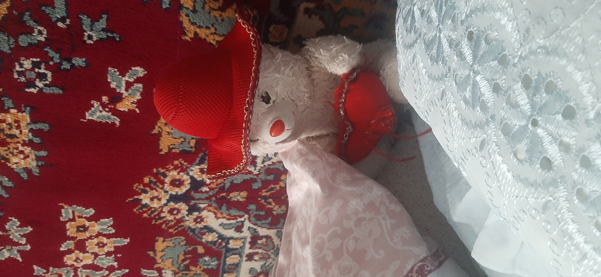 A stuffed bear with a red hat and a red heart is lying on a patterned rug and bedding. The bear is white and fuzzy, with a red nose and dark eyes. It is wearing a red crocheted hat with a gold trim. In its paws, it holds a small red heart, also adorned with gold trim and red string. The bear is positioned on a pink blanket with a floral pattern, which is itself placed on a white, lace-like fabric. The background features a red rug with intricate floral designs in yellow, blue, and white. The scene is indoors, likely in a domestic setting. There is no discernible information in the image to indicate a specific location within Yessentukskaya, Russia.