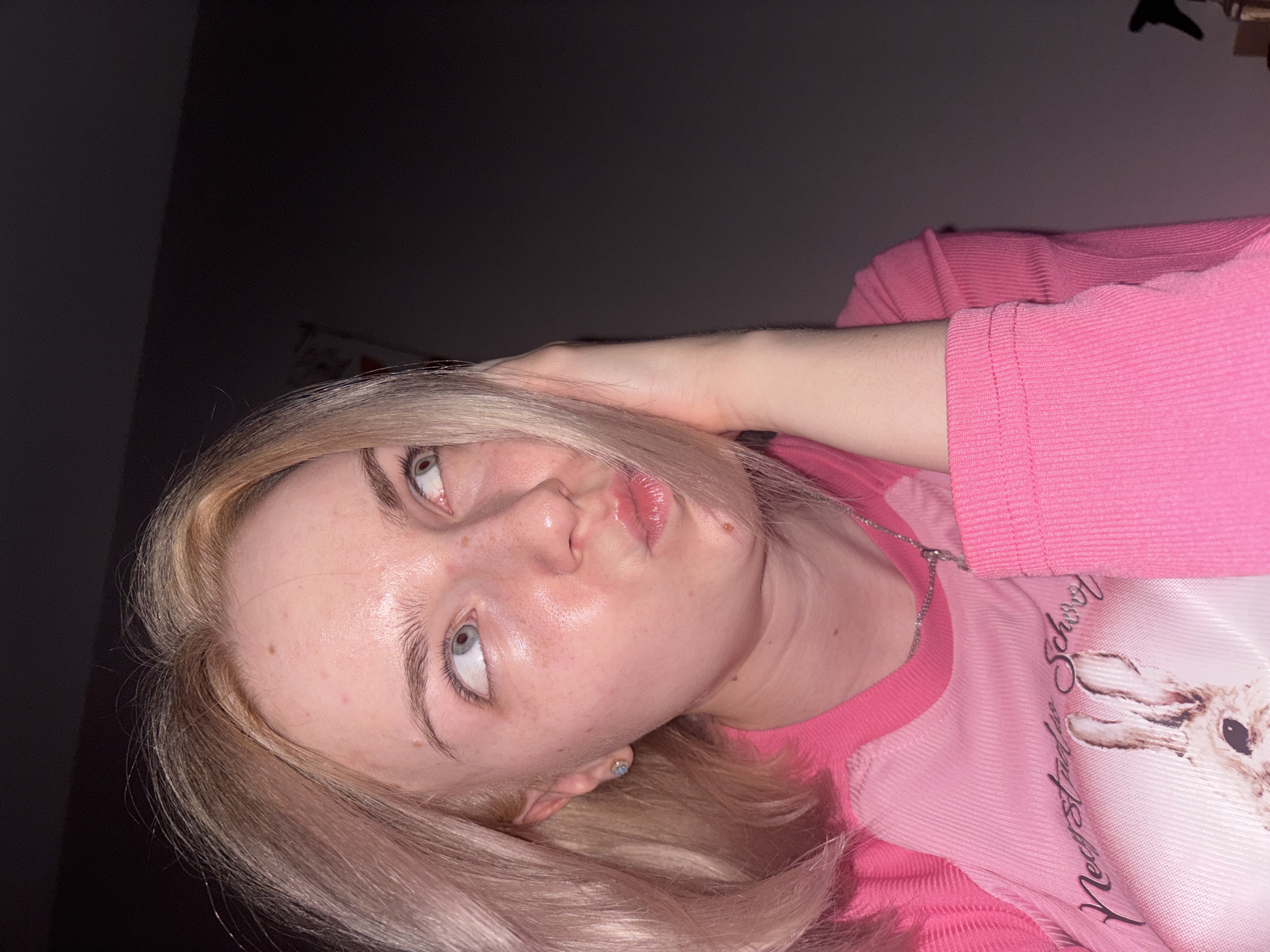 A close-up, tilted portrait shows a young individual, likely female, with blonde hair and light-colored eyes. Her head is angled, and her gaze is directed upwards and slightly to the left, with her lips slightly pursed in what appears to be a contemplative or playfully pouting expression. Her complexion is fair, with some minor skin blemishes visible.
She is wearing a pink, long-sleeved top. The top features a ribbed texture on the sleeves and shoulders, while the main body appears smoother. On the front of her shirt, a graphic design depicts a rabbit's head, and partially visible text reads "New Future School." A silver chain necklace is visible around her neck, and a small, light-colored stud earring is in her left ear.
One of her hands is raised, with fingers gently touching the back of her head or hair, while her forearm is visible against the dark background. The setting appears to be indoors, characterized by dim lighting that suggests it is evening or night. The background is mostly dark and indistinct, with only a very faint outline of an object (possibly a hanger or fixture) visible in the upper right corner. The overall mood is casual and intimate, captured in what appears to be a selfie or a relaxed pose.