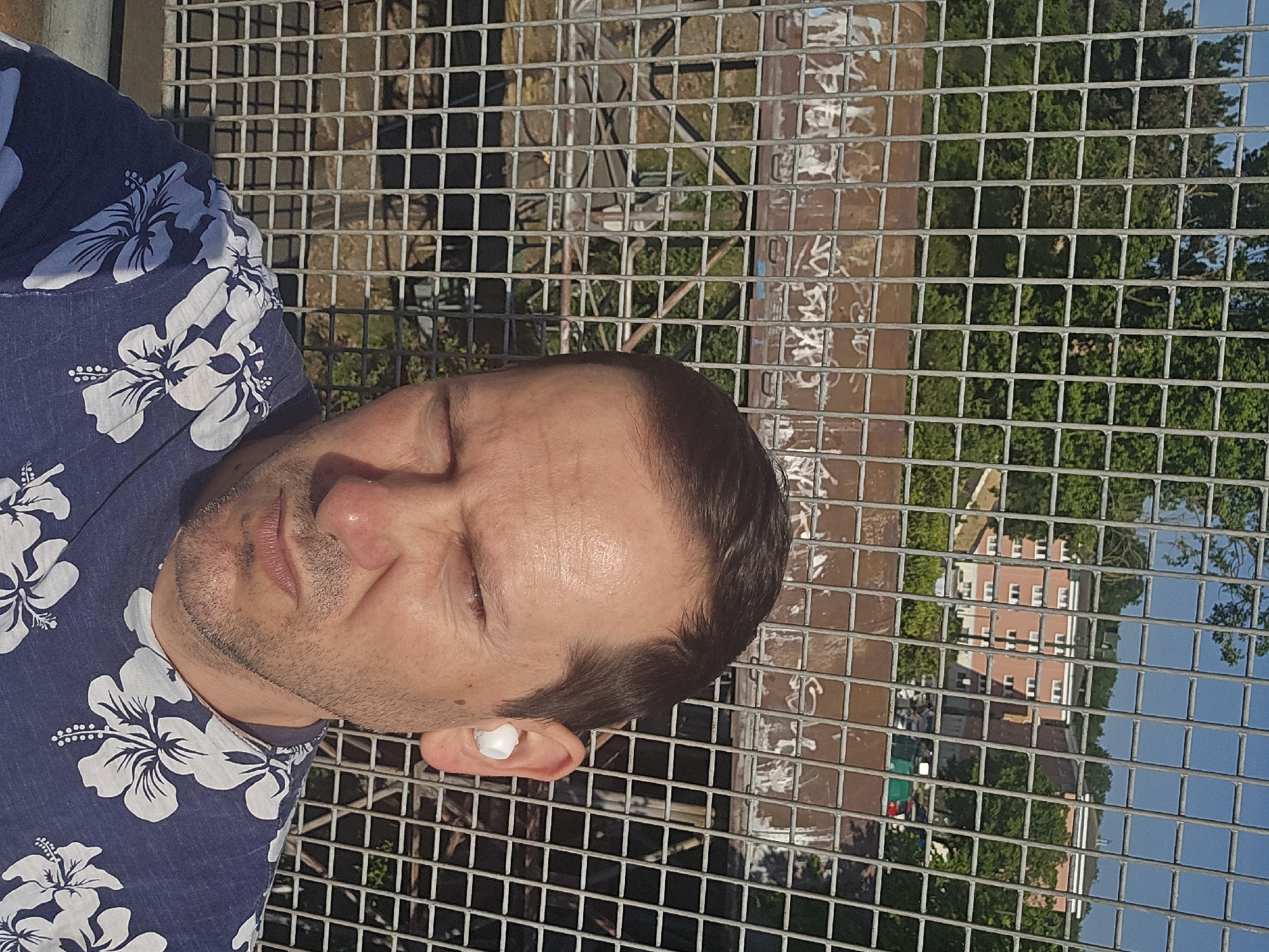 A close-up shot shows a man with his eyes closed, wearing a blue shirt with white floral patterns, positioned against a wire mesh fence. A white object, resembling an earbud, is visible in his ear. The background, seen through the fence, includes buildings, trees, and a portion of the sky. The scene is brightly lit, suggesting it is daytime. The man appears to be at a location overlooking a cityscape or a park. The reflection of a building can be seen in the fence. Without additional information, it's impossible to verify the location in London, United Kingdom.