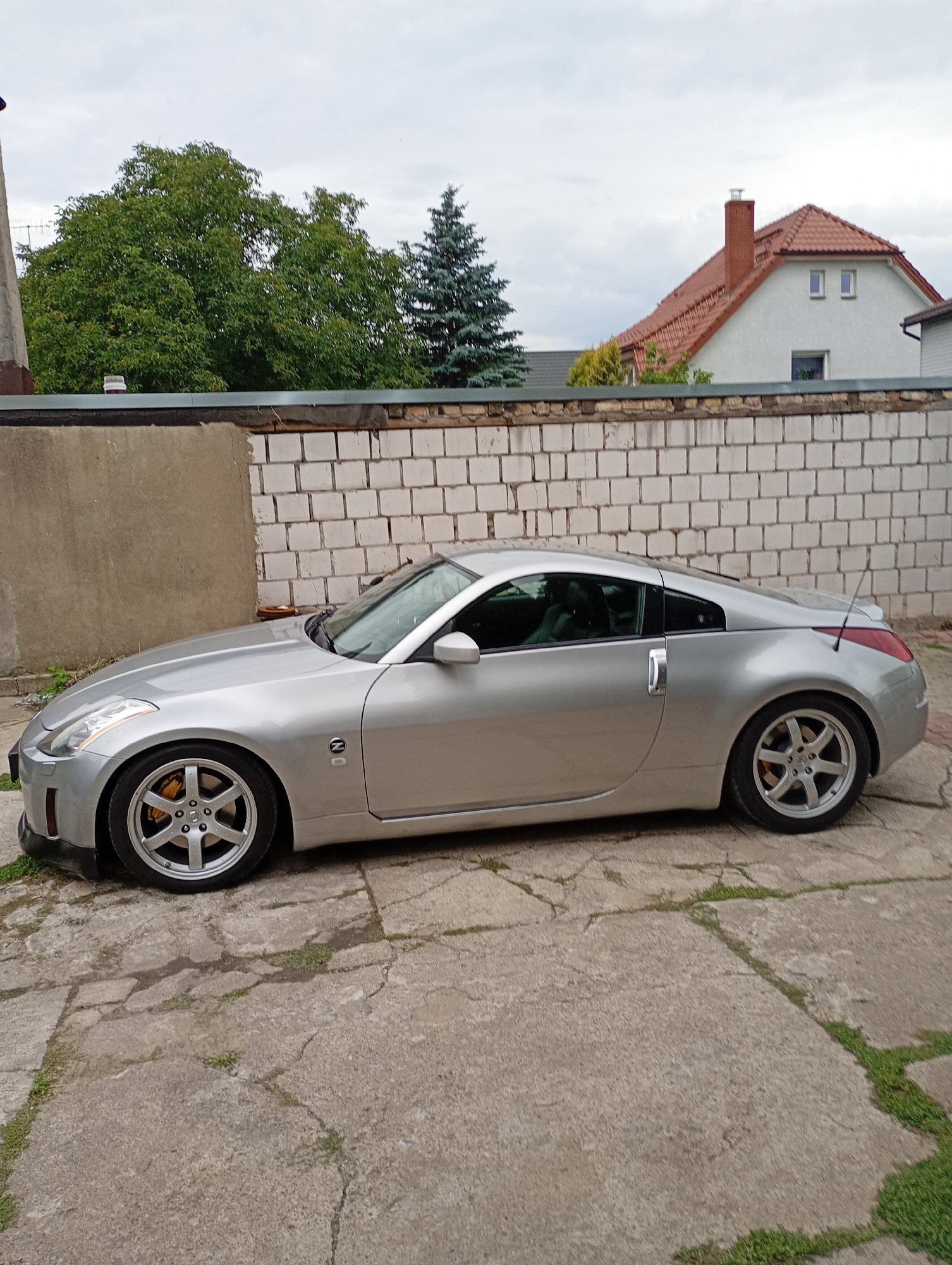 A silver Nissan 350Z is parked on a cracked concrete driveway in front of a white brick wall. Trees and a residential house with a red tiled roof are visible in the background under a cloudy sky. The car is viewed from its side, showcasing its sporty design. The overall scene appears to be a residential backyard.