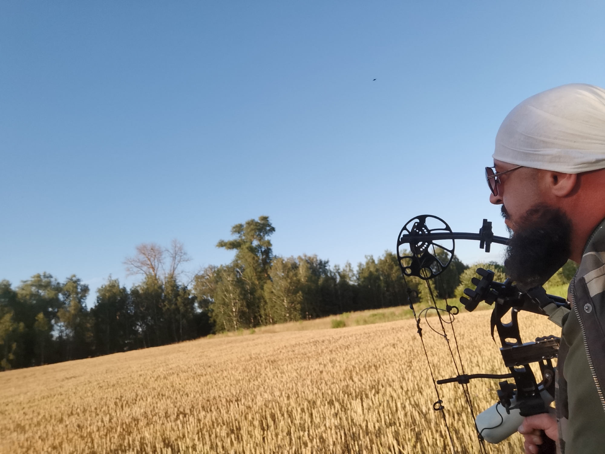 A man wearing a white headscarf and sunglasses is holding a compound bow, aimed towards the right of the frame. He has a beard and is wearing a camouflage jacket. The man is standing in a field of wheat with a treeline in the background. The sky is clear and blue. The scene appears to be set in a rural area. The time of day is likely late afternoon or early evening, given the warm lighting on the wheat field. The weather is clear and sunny. No text is visible in the image.