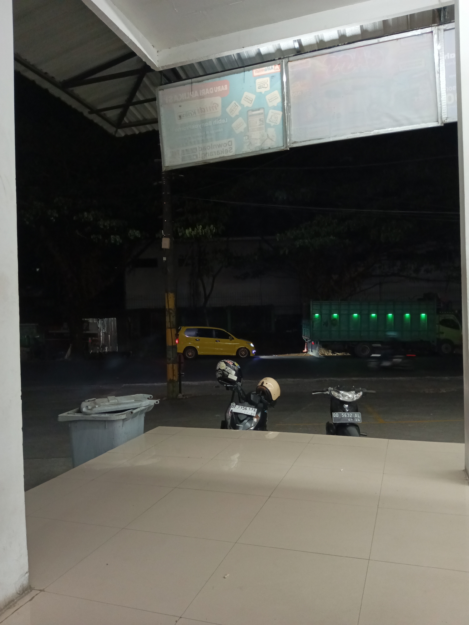 The image depicts an outdoor scene at night, possibly in Makassar, Indonesia. In the foreground, a tiled floor extends towards a street. To the left, a large gray trash bin with an open lid is visible. Further back, two motorcycles are parked. The motorcycle on the left has a white helmet on its seat and the license plate reads "DD 1756 X VK". The motorcycle on the right has a beige helmet on its seat and the license plate reads "DD 5632 AL".
The street features a yellow car parked to the left, and a green truck with illuminated windows is moving to the right. In the background, trees and dark structures are visible under artificial lighting. Two large signs with text and images are mounted overhead. The overall impression is a nighttime urban environment.