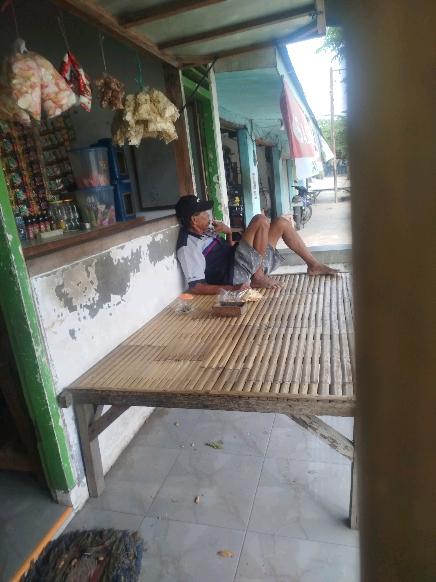 An elderly Indonesian man with a black cap sits cross-legged on a bamboo mat bench outside a roadside stall in Tonjung, Indonesia. He is wearing a blue and white short-sleeved shirt and grey patterned shorts. The stall, made of wood and tin, displays bags of snacks hanging from the ceiling and bottles of drinks on shelves. A motorcycle is parked in the background. The ground is tiled. The scene appears to be a typical rural or suburban Indonesian setting.