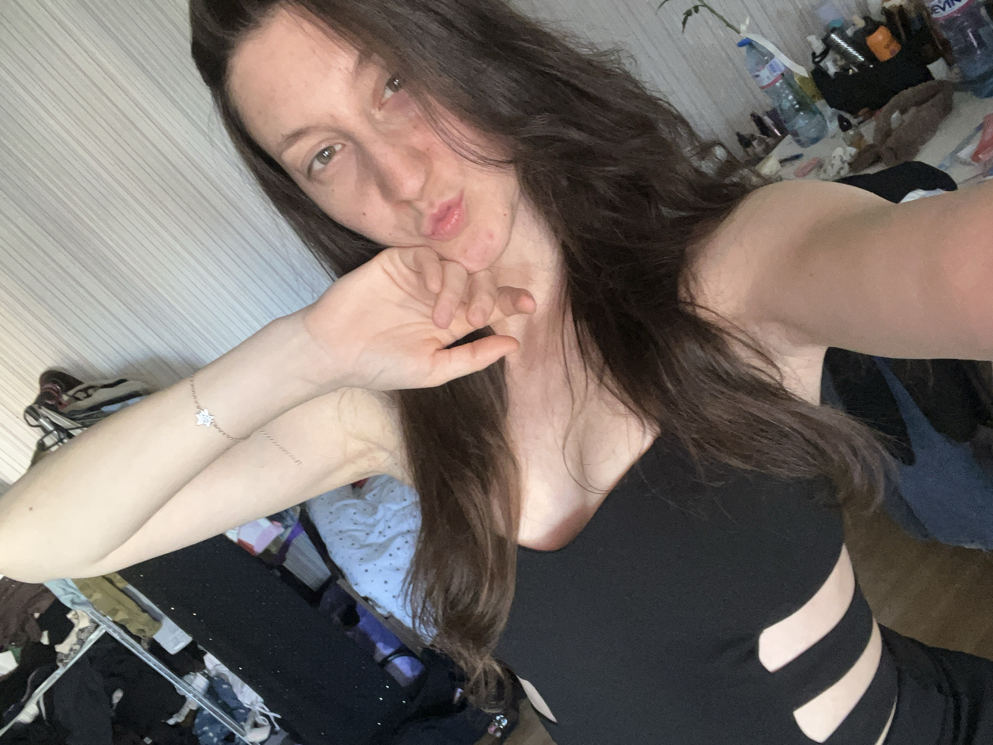 A selfie taken by a woman with long, wavy dark brown hair, fair skin, and facial acne, wearing a black sleeveless top with side cutouts. She has one hand near her face, as if posing, and is looking directly at the camera with a slightly puckered expression. A silver-colored bracelet is visible on her wrist. The background is cluttered, including items like water bottles, clothing, and other personal belongings. The setting appears to be a messy interior, likely a bedroom or living space, in an Unknown location within an Unknown city.