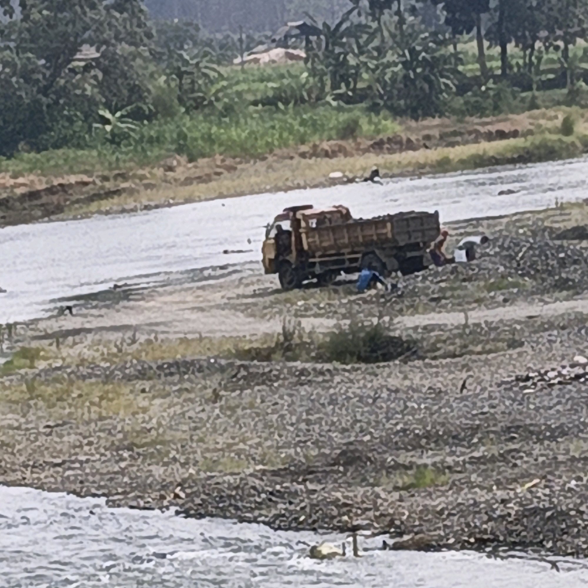 In Purana, Indonesia, a construction site is visible next to a body of water. A yellow dump truck is parked on a gravelly bank, with its bed loaded with dirt or sand. Two individuals are near the truck. One person is partially visible in the driver's seat, while another is kneeling or squatting beside the truck, possibly working. Further in the background, lush green vegetation and trees line the landscape. The water appears to be a river or stream, with some ripples and small waves. The overall scene depicts a work in progress at a riverside location.