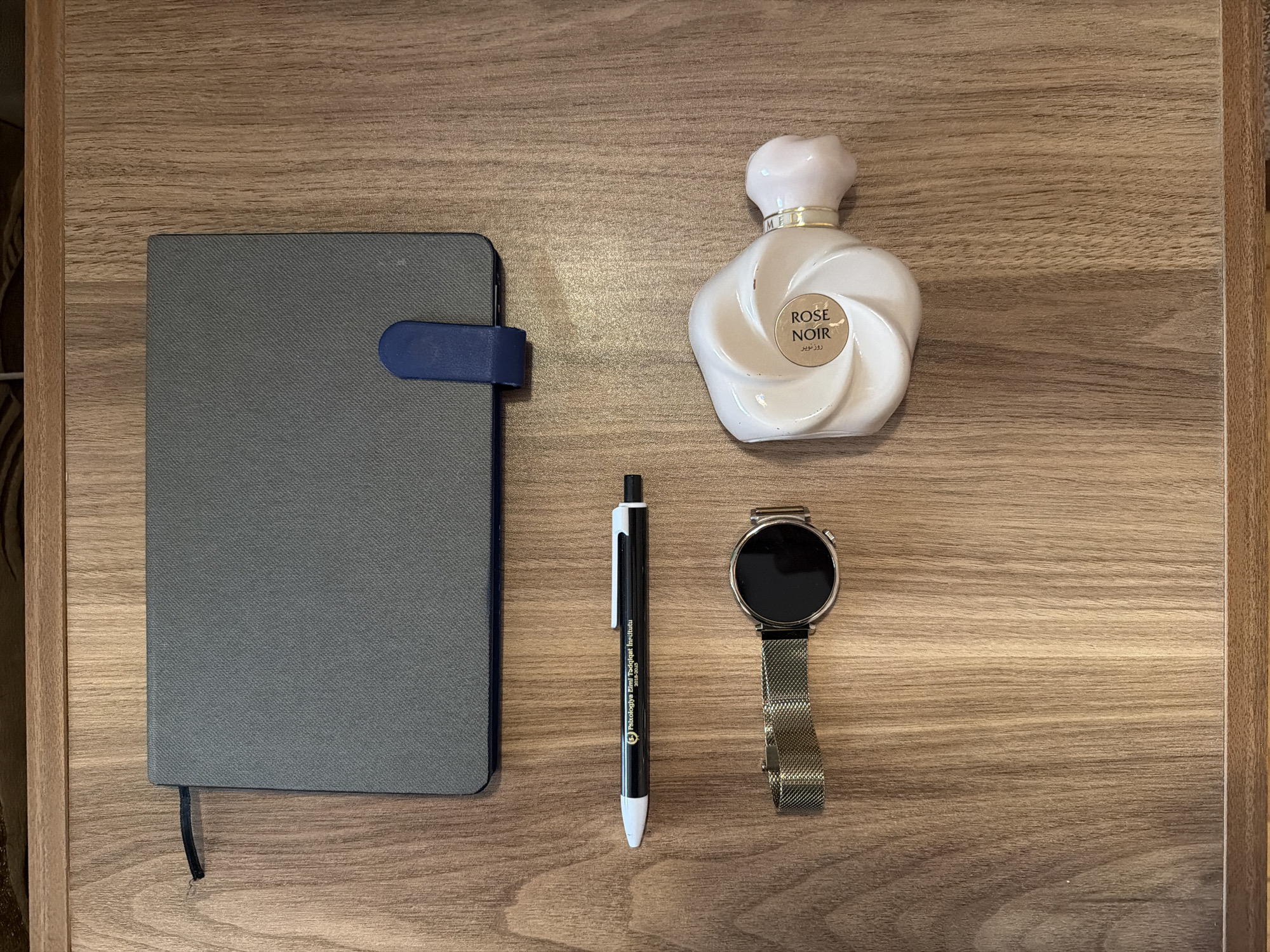 The image displays a flat lay of various personal items on a wooden surface. From top to bottom, there is a grey notebook with a blue elastic band. Next, a black pen with white and gold accents, bearing the text "Psixologiya Elmi Tədqiqqat İnstitutu 2015-2015". Below the pen is a smartwatch with a gold mesh band and a circular black screen. To the right of the smartwatch is a bottle of perfume with a distinctive swirling design, labeled "ROSE NOIR". The wooden surface has a prominent grain pattern. The image does not contain any information or visual cues related to the city of Sabirabad, Azerbaijan.