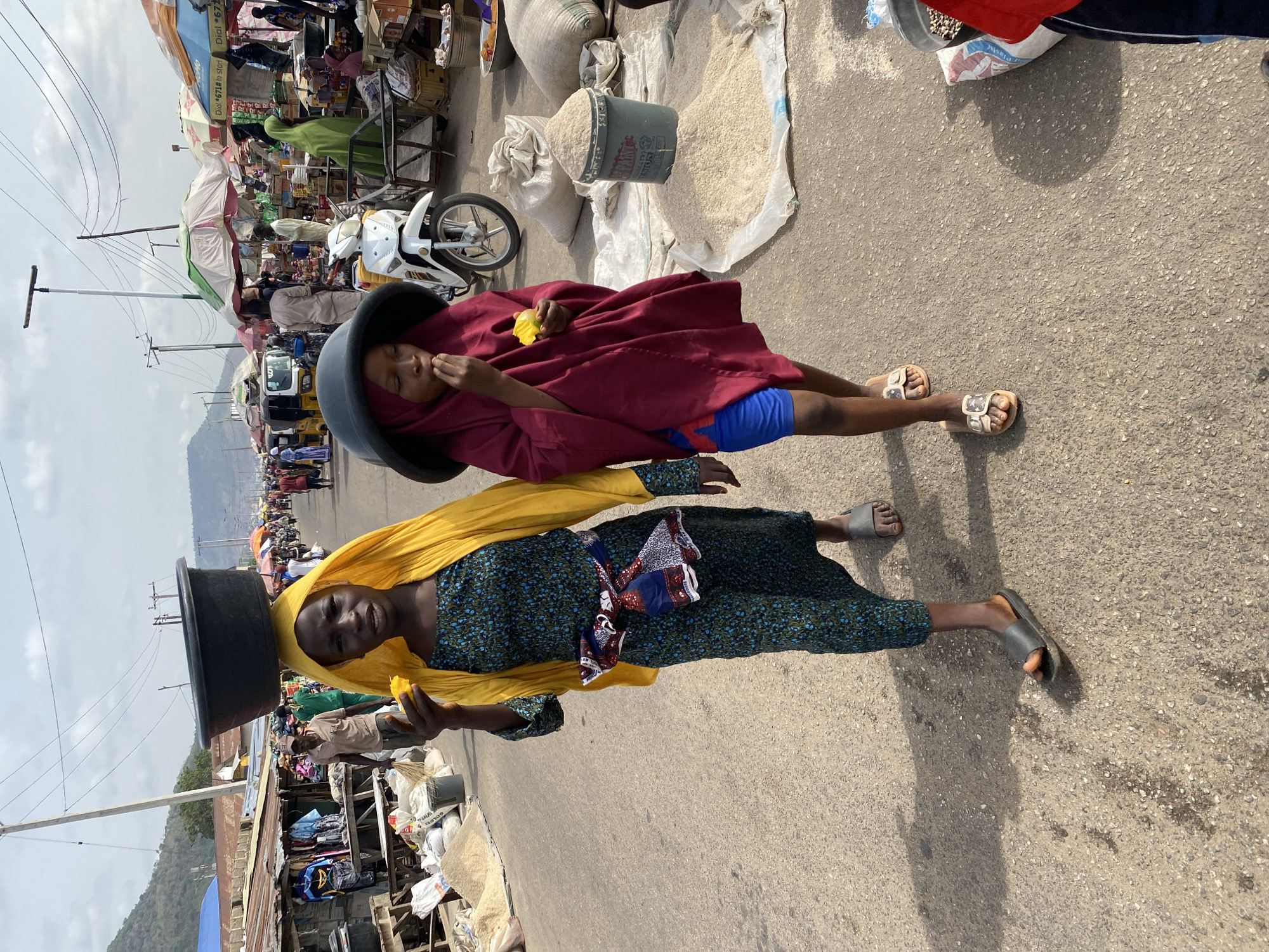Two women are positioned in the middle of a paved road in Lokoja, Nigeria. Each woman balances a large, dark-colored container on her head. The woman in the foreground wears a yellow head covering, a blue and green patterned dress, and dark sandals. She holds a piece of fruit in her hand and looks toward the camera. The second woman is dressed in a maroon-colored robe and blue shorts. She is also holding a piece of fruit. Behind the women is a bustling marketplace with tents, vehicles, and numerous people. Bags of grain and other items are situated near the women. In the background, there are hills visible on the horizon.