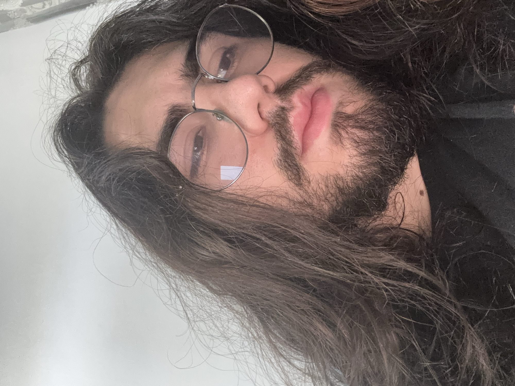 A close-up, low-angle shot captures a person with long, dark, wavy hair and a beard. They are wearing round, thin-rimmed glasses. The person's mouth is slightly open, and their expression is neutral. The background is a plain, light-colored wall, suggesting an indoor setting. The lighting is soft, and there are no discernible objects or features in the environment.