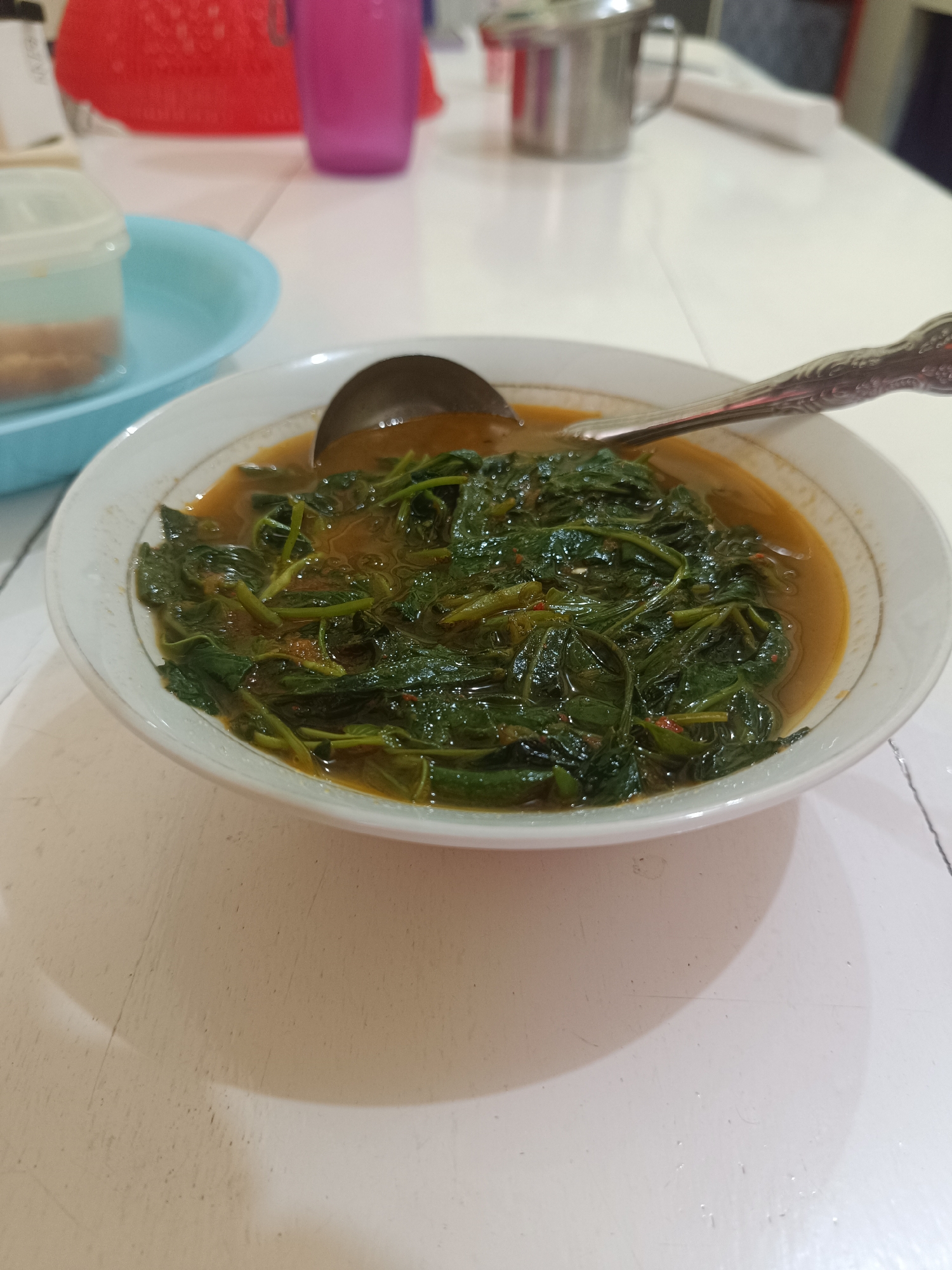 The image displays a close-up view of a bowl of cooked green vegetables in a sauce, positioned on a white table. The central subject is a white ceramic bowl, adorned with a subtle gold rim, brimming with a dish primarily composed of dark green leafy vegetables, likely water spinach (kangkung), immersed in a viscous yellowish-brown sauce. A silver metal ladle with a decorative, ornate handle rests within the bowl, partially submerged.
In the blurred background on the left, a light blue plastic bowl or plate is visible, accompanied by a small, clear plastic container with a white lid, possibly holding spices or condiments. Further behind these items, a vibrant red plastic basket or container is partially discernible. To the right in the background, a magenta or purple plastic cup and a silver metal mug with a handle can be seen.
The scene suggests an indoor setting, such as a dining table in a home or a casual restaurant. The white table surface shows some minor wear and faint marks. The lighting is soft and diffused, characteristic of indoor ambient light, making the precise time of day difficult to ascertain. There are no individuals present, and no visible text in the image. The meal, consistent with Indonesian cuisine from the Lhokseumawe region, appears to be a ready-to-serve dish.