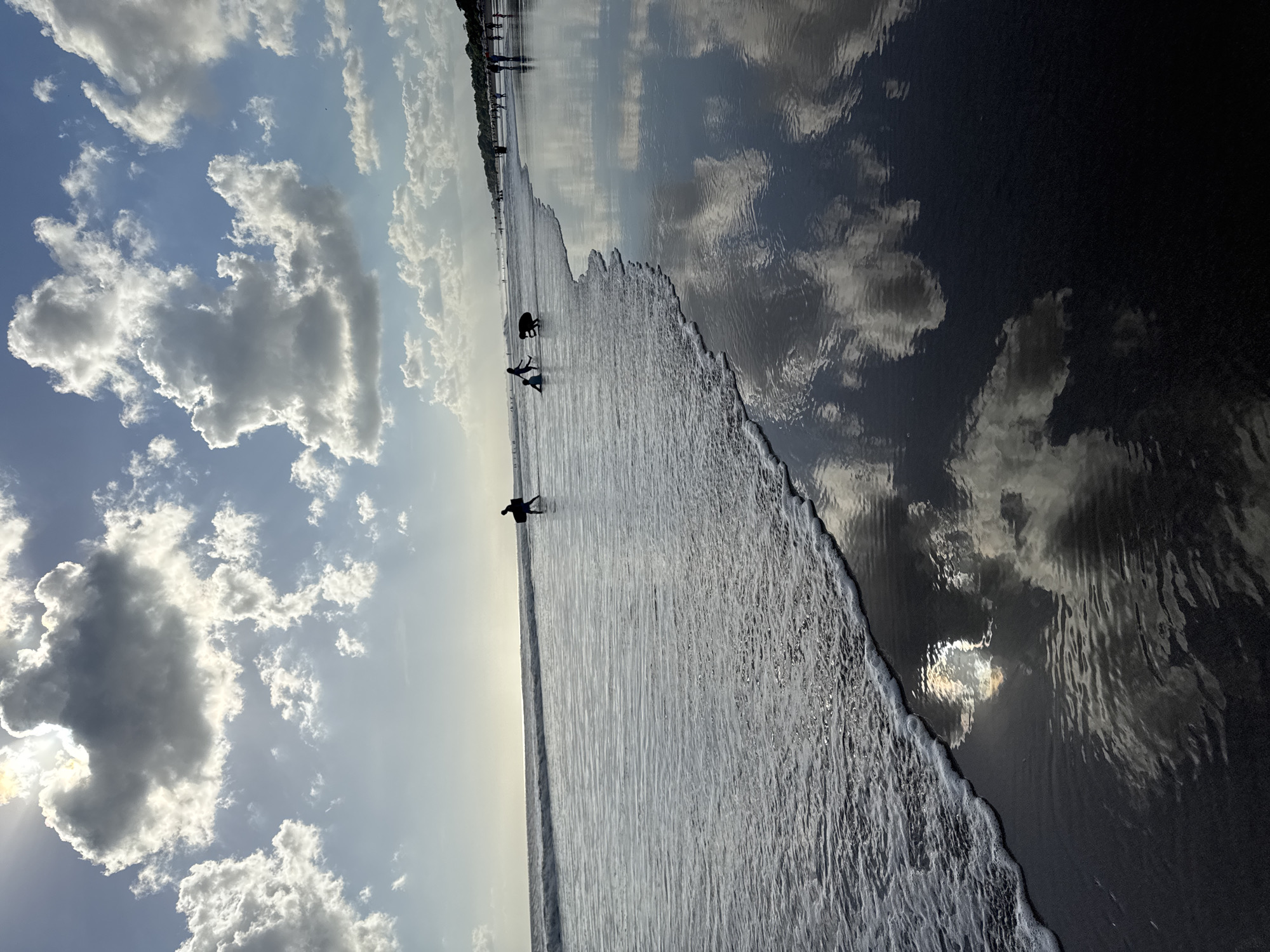 The image depicts a beach scene in Kuta, Indonesia, with a focus on the shoreline and the interplay of light and water. The sky is filled with dynamic clouds, some brightly lit by the sun and others casting shadows. The sun itself is partially obscured by a large, dark cloud, creating a dramatic lighting effect.
In the foreground, a gentle wave is washing ashore, leaving a thin line of white foam on the dark, wet sand. The wet sand acts as a mirror, reflecting the sky and the clouds with striking clarity. The reflections create a sense of depth and movement in the lower half of the image.
Further out in the ocean, several silhouetted figures can be seen. One individual is walking along the water's edge carrying a surfboard. Closer to the shore, two children are playing in the shallow water. In the distance, more people are walking along the beach, their figures also appearing as silhouettes against the brighter horizon. The overall atmosphere is one of natural beauty and leisure activity.