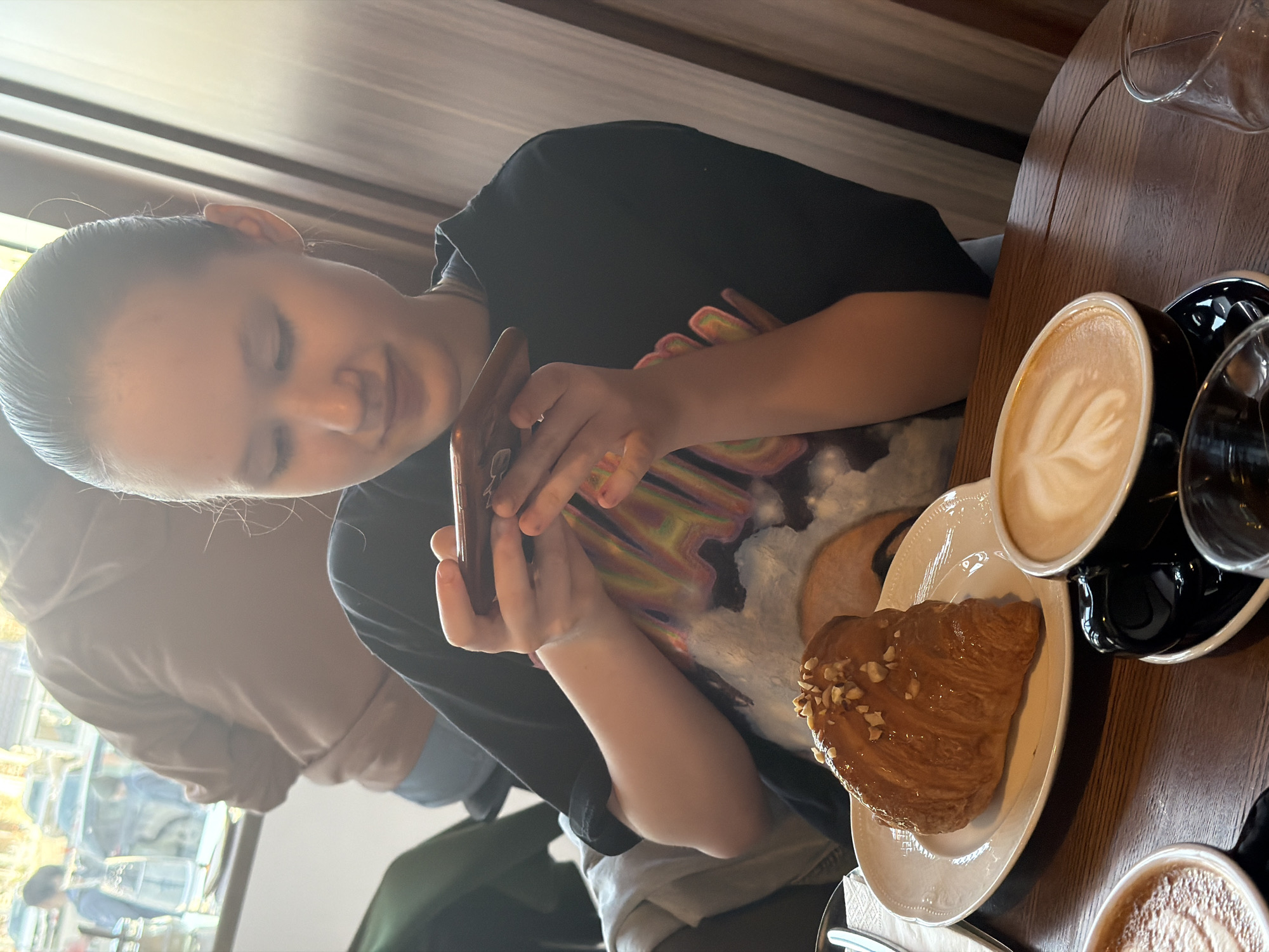 A young woman sits at a wooden table in what appears to be a café. She is holding a brown smartphone in her hands, focusing her attention on the device. She is wearing a black t-shirt with a colorful graphic print. On the table is a white plate holding a croissant with sliced nuts. Adjacent to the plate is a black cup of coffee with latte art and a clear glass of water. Another cup with coffee is also visible at the bottom of the frame. The café appears to have a modern interior with wood paneling on the wall. Through the window, a city street with other people and buildings is visible. The location is identified as Perm, Russia.
