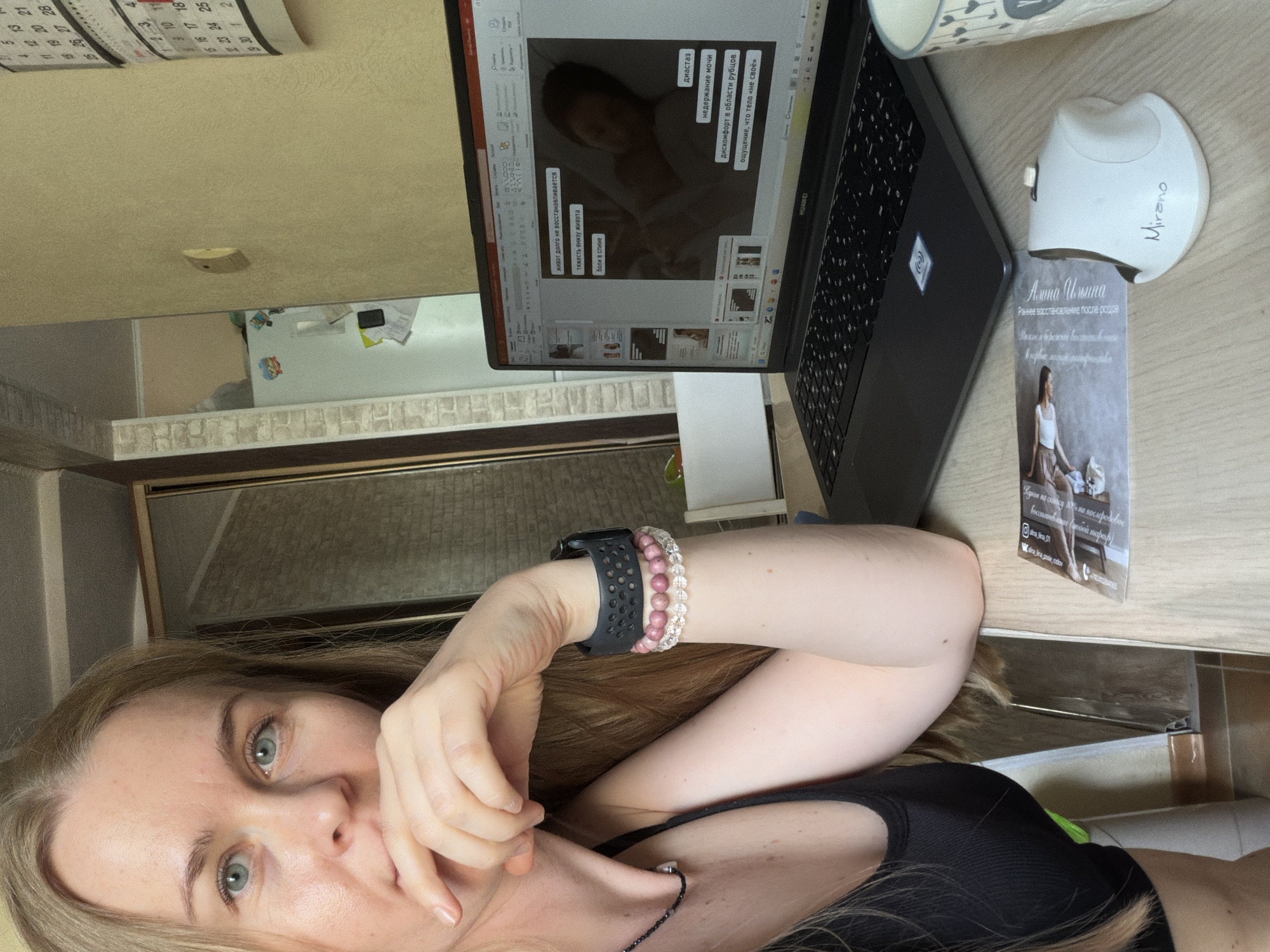 A woman with blonde hair and blue eyes is sitting at a wooden desk in what appears to be a home office. She is wearing a black tank top and several bracelets on her left wrist. She is looking towards the camera with her hand on her chin. On the desk in front of her is an open laptop displaying a presentation slide with text in Russian and an image of a woman lying in bed. To the right of the laptop is a white computer mouse and a cup with a heart pattern. A calendar hangs on the wall behind her, indicating dates in May and June. The room has light yellow walls and a brick accent wall to the left. The overall environment suggests a casual, indoor setting. There is no information in the image to suggest it is located in Khabarovsk, Russia.