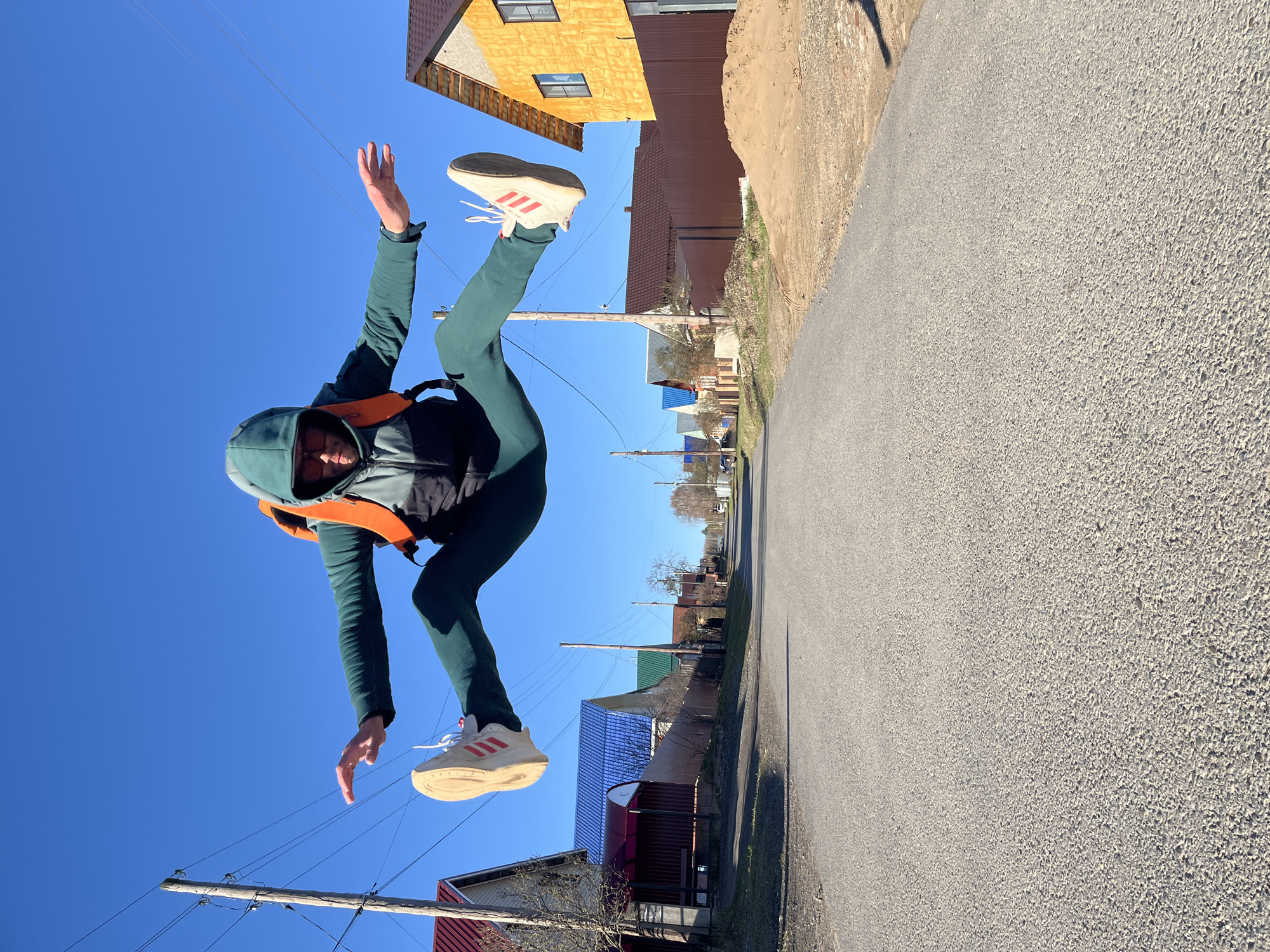 The image captures a person in mid-air, performing a jump or acrobatic pose on a bright, sunny day. The individual wears a dark green hooded jacket, a black vest with an orange strap, dark green pants, and white sneakers with red stripes, characteristic of Adidas. One arm is raised, the other lowered, and one leg is bent while the other is extended, creating a dynamic, energetic impression.
The background features a clear, deep blue sky. Below the person is an asphalt road, upon which strong, distinct shadows are cast, indicating bright sunlight, likely during midday or early afternoon. Along the side of the road, various residential houses are visible, featuring details like yellow siding, brown fencing, and roofs in shades of blue and red. Wooden utility poles with overhead wires run through the scene. The setting suggests an outdoor street or residential area in Chaikovsky, Russia. No specific text is clearly legible on any structures, though the branding on the shoes is implied by the stripe pattern.