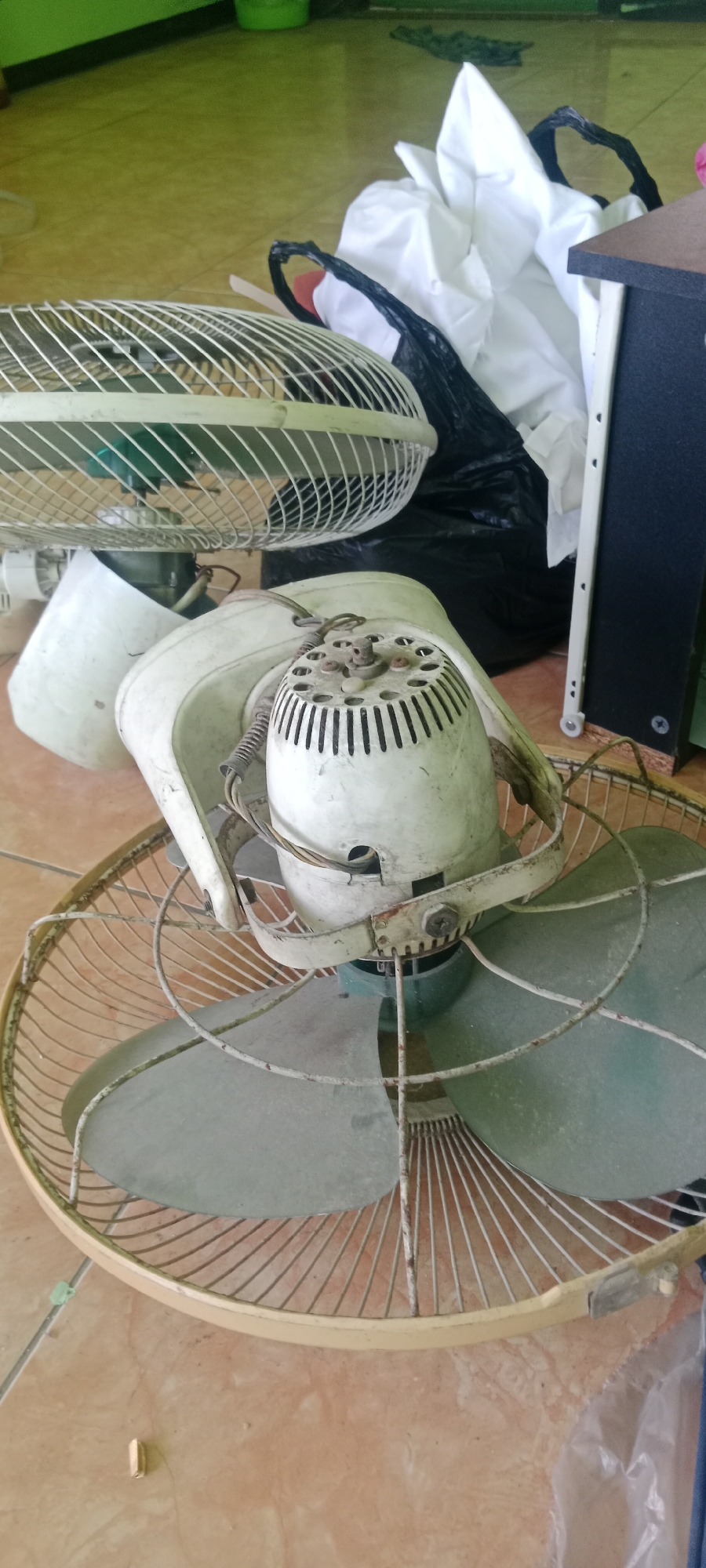 The image shows a close-up of several old, dusty electric fans on a tiled floor. The fans appear to be disassembled or broken, with exposed wires and discolored plastic parts. In the background, a black plastic bag filled with white fabric is visible, along with a portion of a dark-colored piece of furniture or equipment. The overall impression is one of disarray and neglect. The flooring suggests an indoor setting, possibly a storage area or a room where items are being repaired or discarded. Without further context or identifying landmarks, it is not possible to definitively place this scene within the city of Tuban, Indonesia.