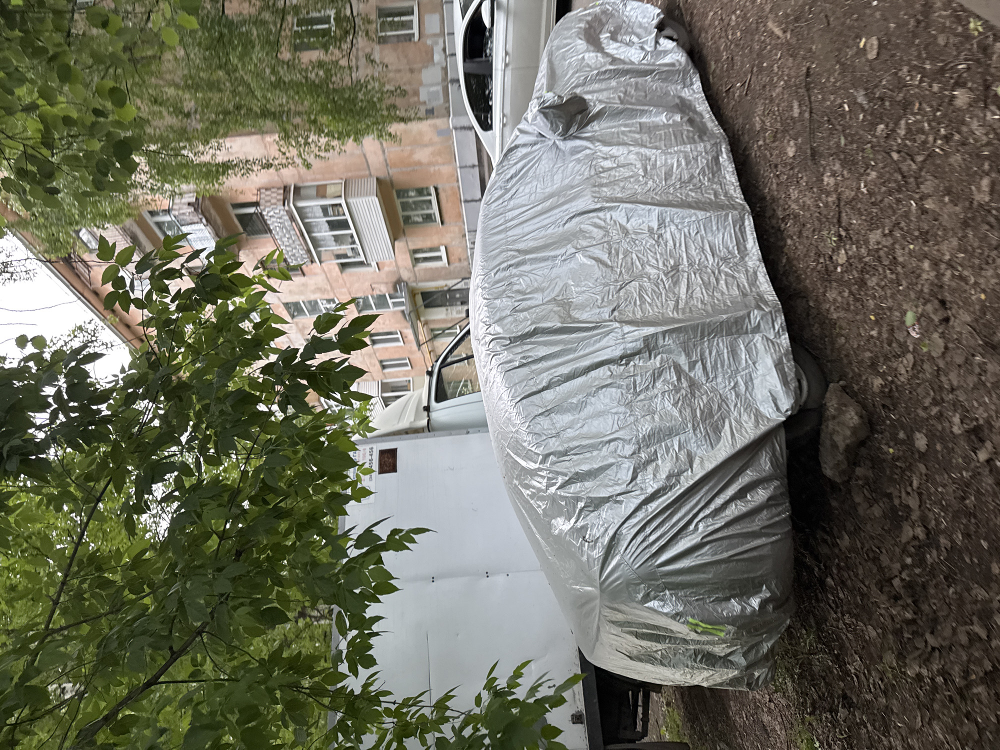 The image depicts an outdoor urban scene in Izhevsk, Russia, characterized by an overcast sky and lush green foliage. In the foreground, a vehicle is entirely covered by a silver, crinkled protective cover, parked on dark brown, uneven earth with scattered small rocks and debris. A bright green or yellow strap is visible on the left side of the car cover.
To the immediate left and partially behind the covered car, a large white box truck is visible, with its cargo area and a portion of the cab. The truck's side panel features a brown rectangular sticker and white text, including the letters "TA" and numbers "456-456". Dense green tree foliage from deciduous trees in full leaf overhangs the left and upper portions of the scene, partially obscuring the truck and the sky.
In the midground, a multi-story residential building dominates the right side. The building is constructed with light peach or pink-colored facades and features numerous windows with white frames, as well as several balconies, some with metal railings and others enclosed. To the right of this building, another tree with lighter, possibly weeping green foliage is visible. A white sedan is parked behind the covered car, partially visible, and appears to be on a paved surface or road closer to the building. No individuals are visible in the scene, and no specific actions or interactions are occurring. The environment suggests a typical residential area or courtyard.