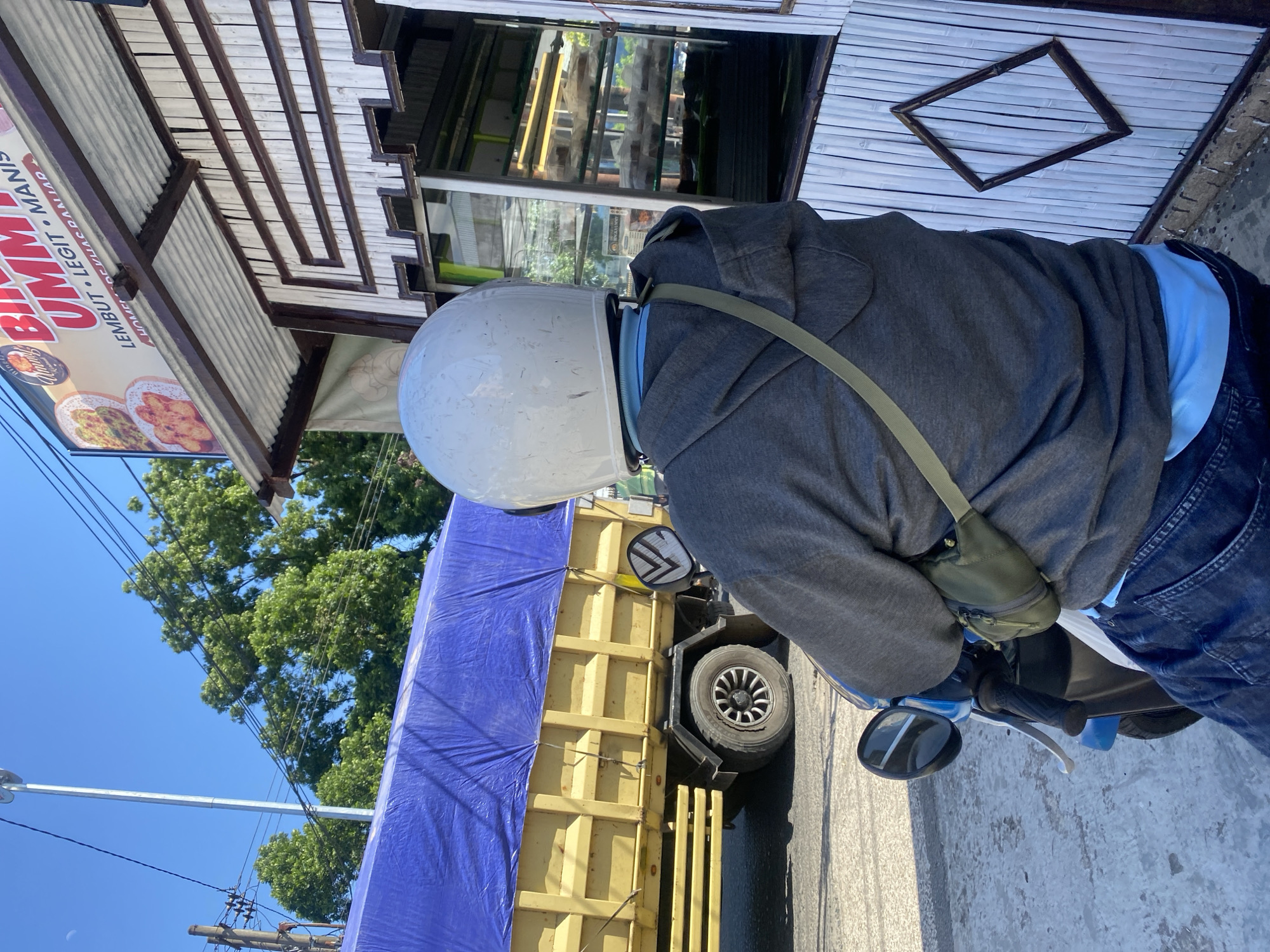A person wearing a white helmet and a grey hoodie sits on a blue and white motorcycle, facing away from the camera. They are positioned next to a small, open-air food stall constructed with bamboo siding and a corrugated metal roof. A sign above the stall reads "BIM UMUM LEMBUT LEGIT MANIS." In the background, a large yellow truck covered with a blue tarp is parked on the side of a street. Trees and utility poles are visible behind the truck. The scene appears to be outdoors, with a clear blue sky overhead. The location is identified as Sungai Paring, Indonesia.